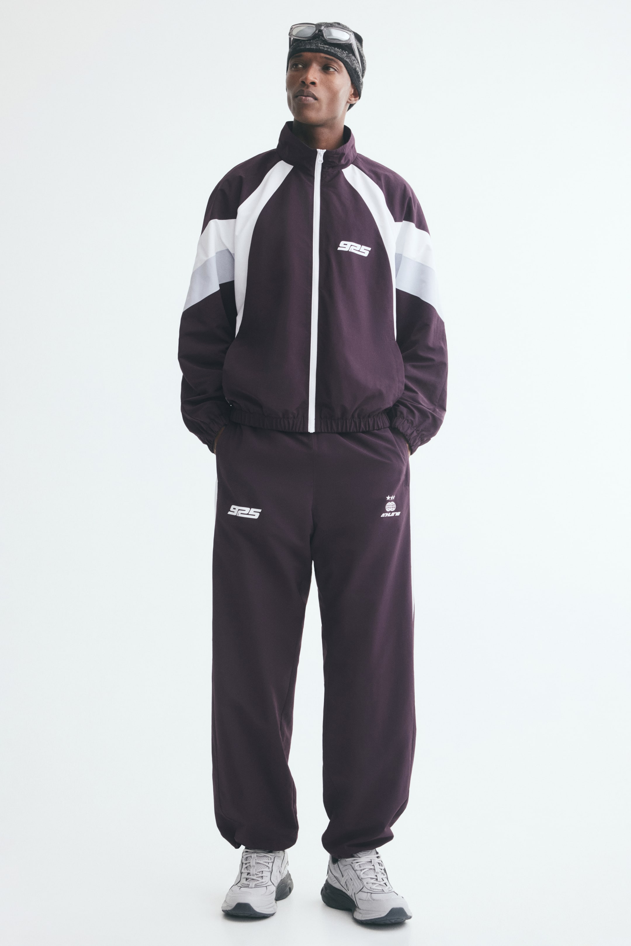 View larger image: Relaxed-fit track pants in dark purple are worn by the model, featuring white printed motifs on the front thighs and elasticized hems. The model is standing with hands resting in the side pockets, styling the pants with a matching color-blocked track jacket and gray sneakers.