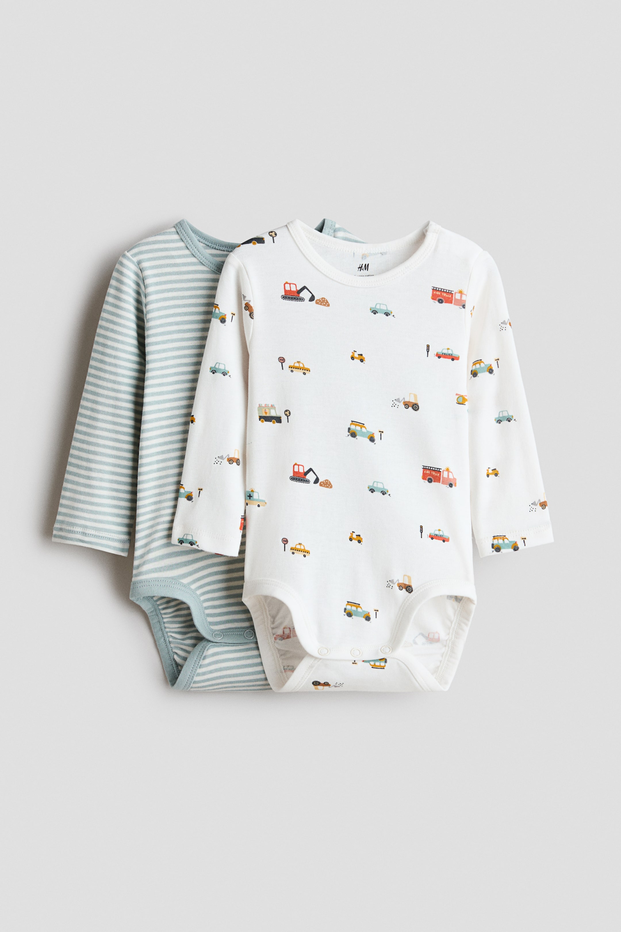 View larger image: Two long-sleeved bodysuits made from soft cotton jersey are showcased from the front. The front bodysuit is white, adorned with a vibrant pattern of vehicles, while the bodysuit layered behind it is light blue with white horizontal stripes. Both bodysuits feature a round neckline, a concealed press-stud fastening on one shoulder, and press-studs at the bottom.