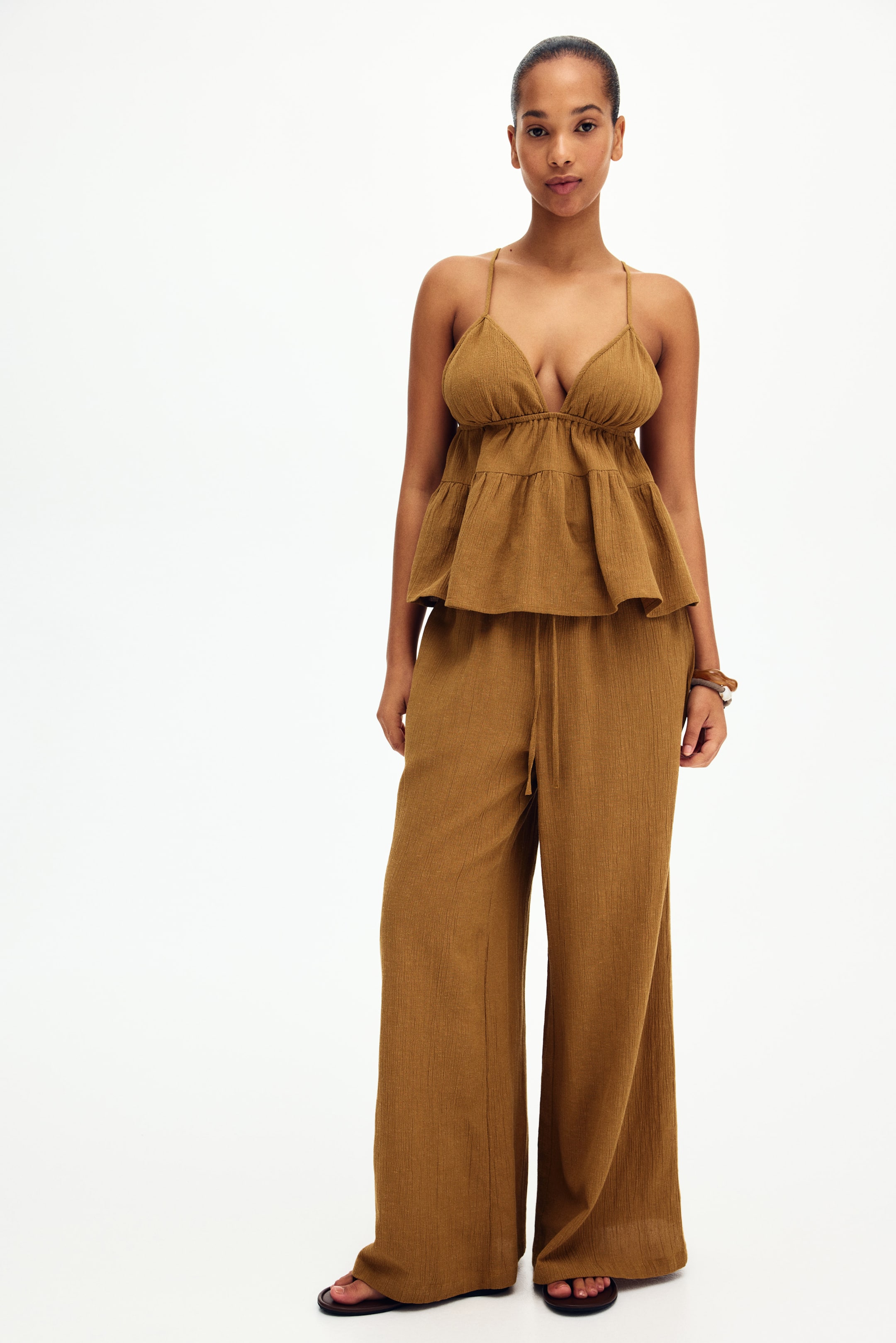 View larger image: Presenting a stylish two-piece set that includes a camisole top crafted in crinkled brown fabric. This top features thin straps, a deep V-neckline, a shirred bust, and a tiered peplum hem for a chic look. Complementing the top are wide-leg pants with a convenient drawstring waist, completing this fashionable ensemble.