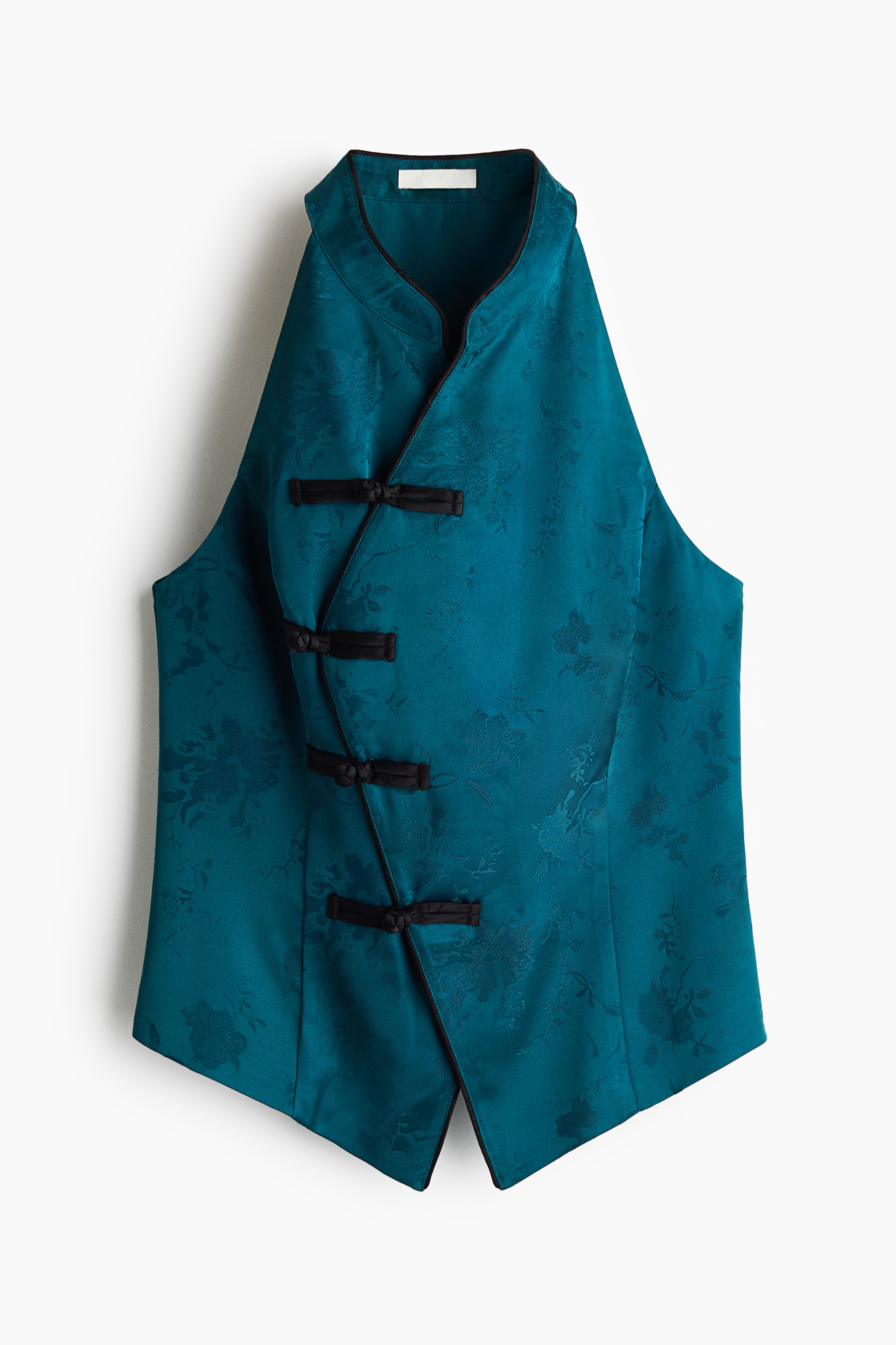 A modell egy ujjatlan, testhezÃ¡llÃ³, tÃ¼rkizkÃ©k felsÅt visel virÃ¡gmintÃ¡s jacquard szÃ¶vÃ©sben. A felsÅ gallÃ©rja Ã¡llÃ³gallÃ©r, nyakrÃ©sze halter stÃ­lusÃº, az aszimmetrikus elÃ¼lsÅ nyÃ­lÃ¡st pedig fekete paszpÃ³lozÃ¡s Ã©s hÃ¡rom csomÃ³zott gombos rÃ¶gzÃ­tÃ©s dÃ­szÃ­ti.