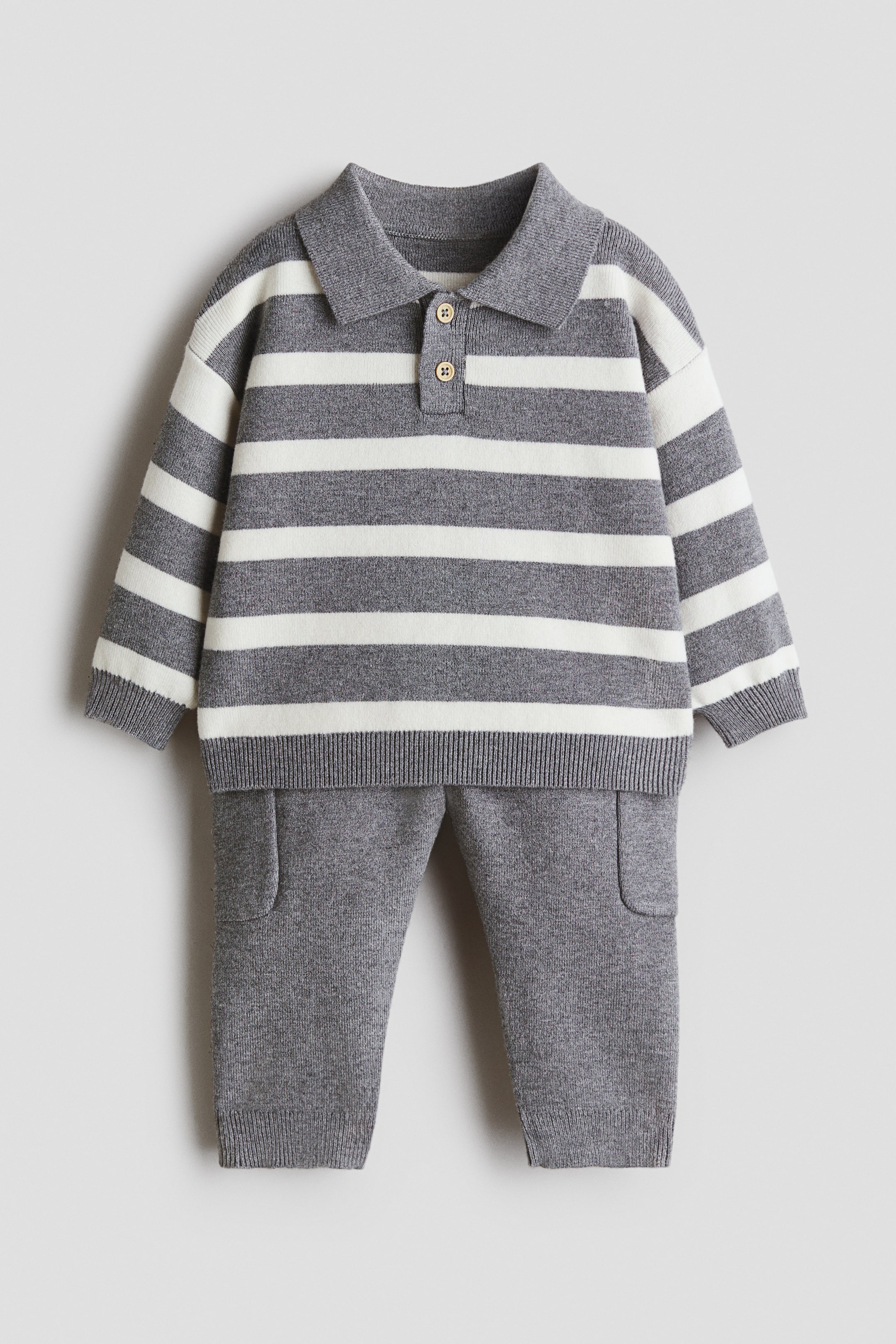 View larger image: A knitted two-piece set is being presented, featuring a grey and white horizontally striped polo sweater with a two-button placket, ribbed cuffs, and a ribbed hem. Matching solid grey knitted pants with patch pockets and ribbed cuffs complete the set.