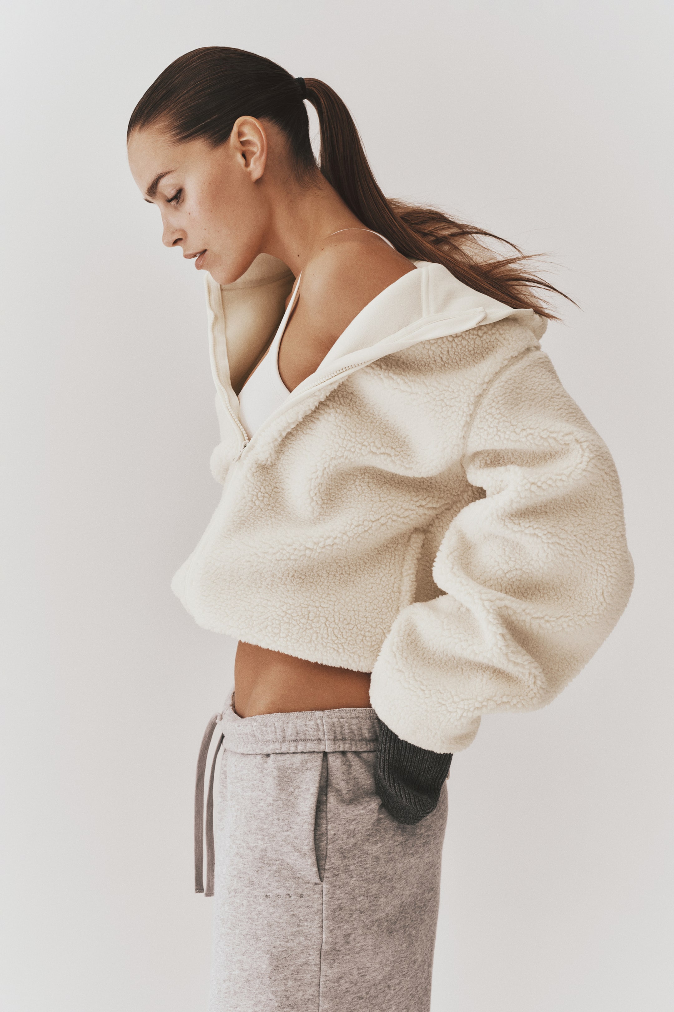 Visa större bild: En teddyfleecejacka i krämfärg med huva och dragkedja framtill bärs av modellen över en vit sport-bh. Den löst sittande, croppade jackan har långa, voluminösa ärmar och stylas med ljusgrå sweatpants.