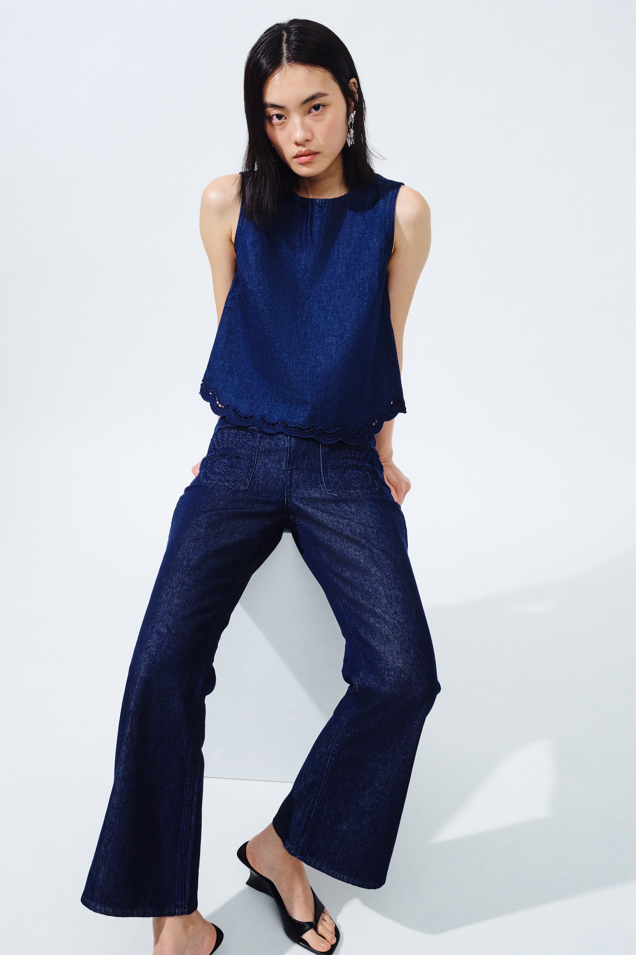 View larger image: A dark blue denim sleeveless top dan seluar lebar yang sepadan dipersembahkan dari hadapan. Top ini menampilkan leher bulat dan hem berombak dengan perincian eyelet, manakala seluar mempamerkan poket tampal depan dengan sulaman yang rumit dan siluet kembang.
