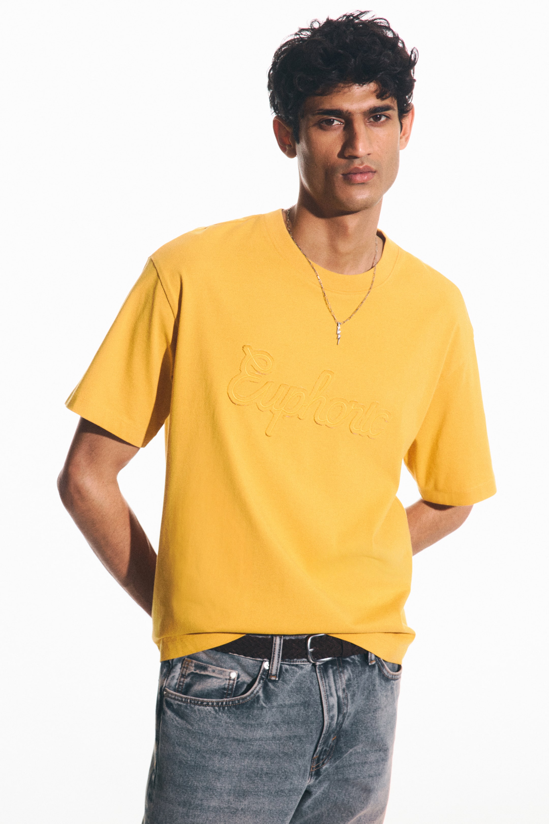 View larger image: A yellow midweight cotton jersey T-shirt, featuring an embossed "Euphoric" text on the front, is being worn by the model. This T-shirt has a round, rib-trimmed neckline, dropped shoulders, a straight-cut hem, and a loose fit, with a silver-toned necklace resting on it.