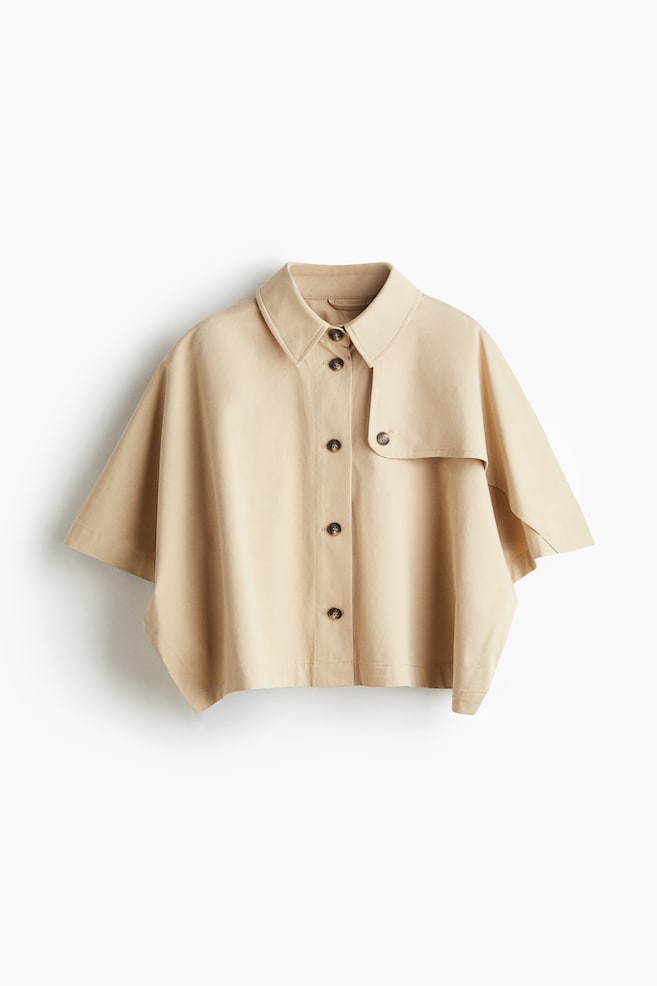 A beige short-sleeved shirt is presented from the front, featuring a pointed collar and a full button-down placket with dark buttons. A storm flap is visible on the right shoulder, secured by a button, and wide, short sleeves are extending from the body. - 1