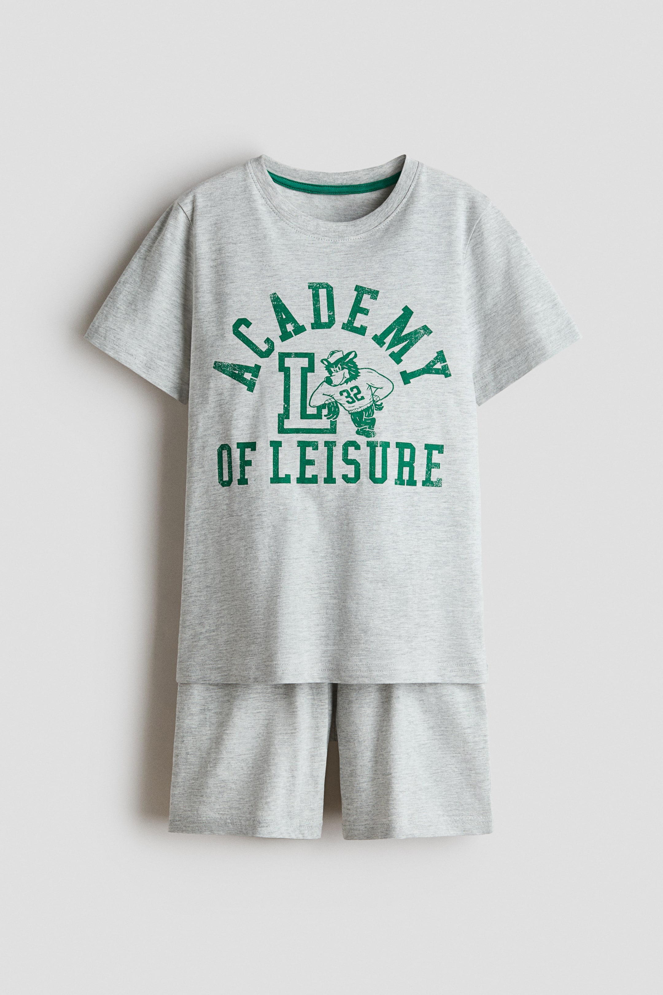 View larger image: Pyjamas in grey marl soft cotton jersey are showcased from the front, comprising a t-shirt and shorts. The t-shirt features a round neckline with green rib-trimming and gently dropped shoulders; its front displays a distressed green 'ACADEMY OF LEISURE' print with a cartoon badger-like character wearing a hat, holding a ball, and numbered 32. The matching shorts come with an elasticated waist.