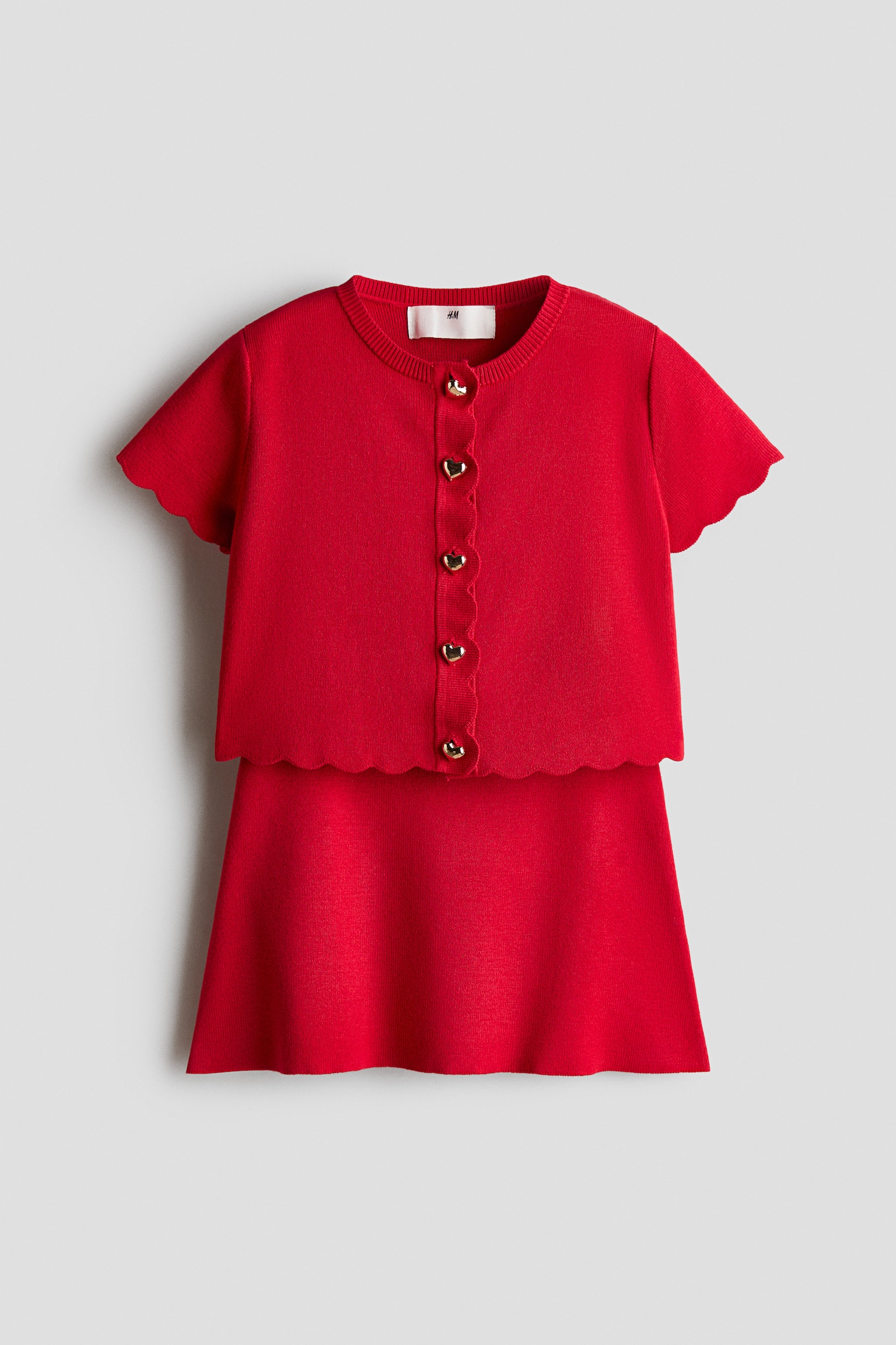 A red knit two-piece set shown from the front features a short-sleeve cardigan with scalloped edges and gold, heart-shaped buttons layered over a matching A-line skirt. The cardigan is showcasing a ribbed crew neck along with scalloped detailing on the sleeves, front placket, and bottom hem.