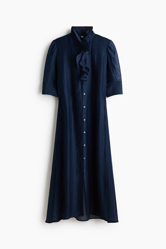 A navy blue midi dress is hanging front-facing, featuring a high collar with cascading ruffles at the neckline and a button-down front closure. The dress is designed with short sleeves with wide cuffs and is crafted from a lightweight, textured fabric with thin ties at the waist. - 1