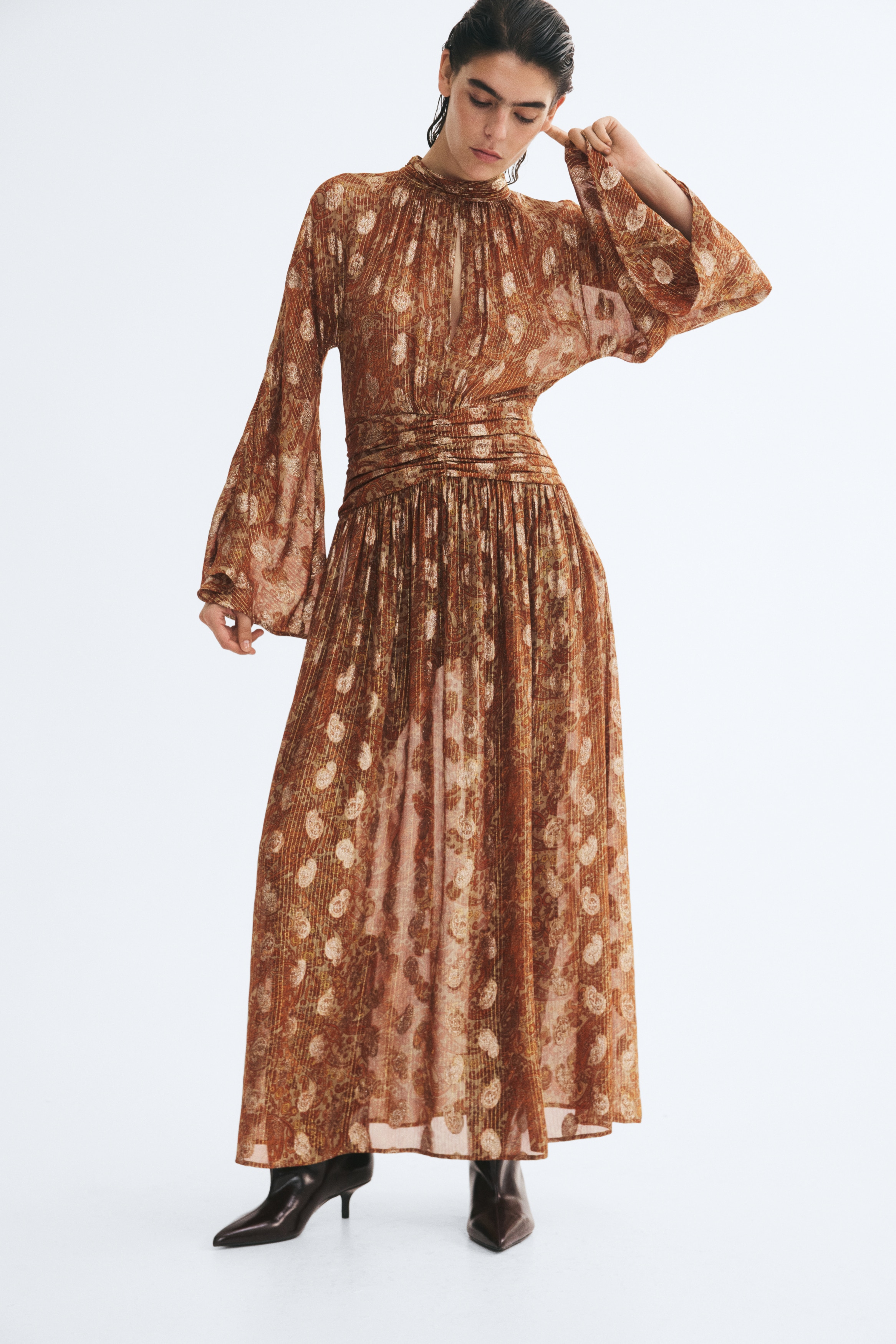 A bronze-colored midi dress facing front features a high band collar with a front keyhole cutout, wide flared sleeves, and a ruched, gathered waistband. The dress displays an all-over paisley pattern with shimmering metallic vertical threads and flows into a pleated skirt.