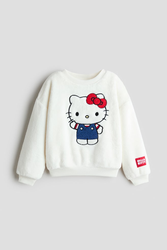 Ein cremefarbener PlÃ¼sch-Sweatshirt ist von vorne zu sehen und zeigt auf der Brustmitte ein groÃes gesticktes Hello Kitty-Motiv. Der Sweatshirt verfÃ¼gt Ã¼ber einen gerippten Rundhalsausschnitt, Ã¼berschnittene Schultern, gerippte BÃ¼ndchen, einen gerippten Saum und einen kleinen roten AufnÃ¤her am linken Ãrmel. - 1