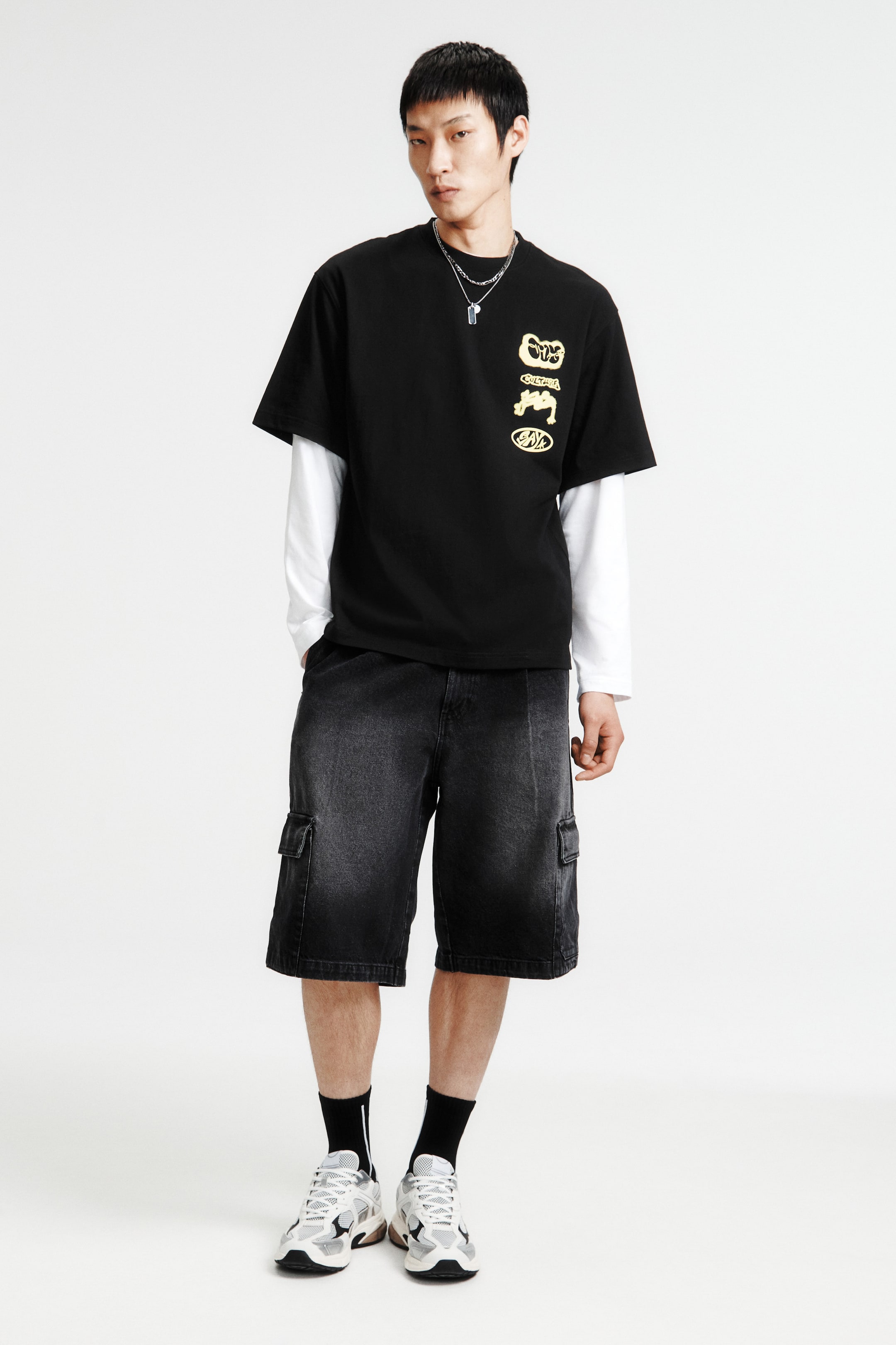 拡大画像を表示: 黒の半袖Tシャツに黄色のグラフィックデザインが施され、白の長袖シャツの上にレイヤードされています。ウォッシュ加工された黒のデニムカーゴショーツと組み合わせられ、サイドポケットがあり、膝まで届く長さです。白とグレーのスニーカー、黒のクルーソックス、シルバーチェーンネックレスがコーディネートを完成させ、正面からのアングルで紹介されています。