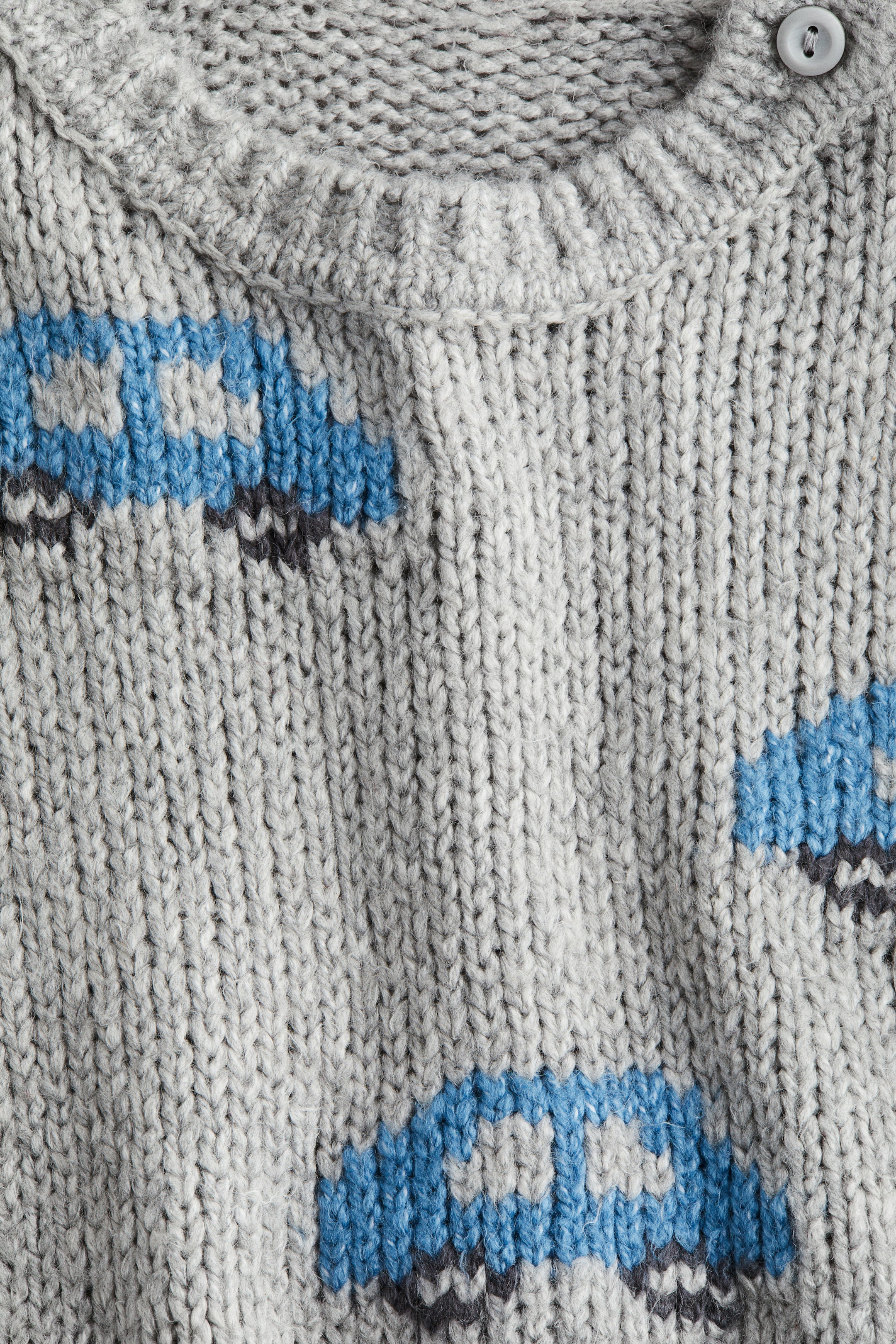 Se større billede: Et gråt, chunky strikket tøj viser et Fair Isle-mønster af stiliserede blå og grå biler. Det har en ribbet krave og en enkelt grå knaplukning nær skulderen.