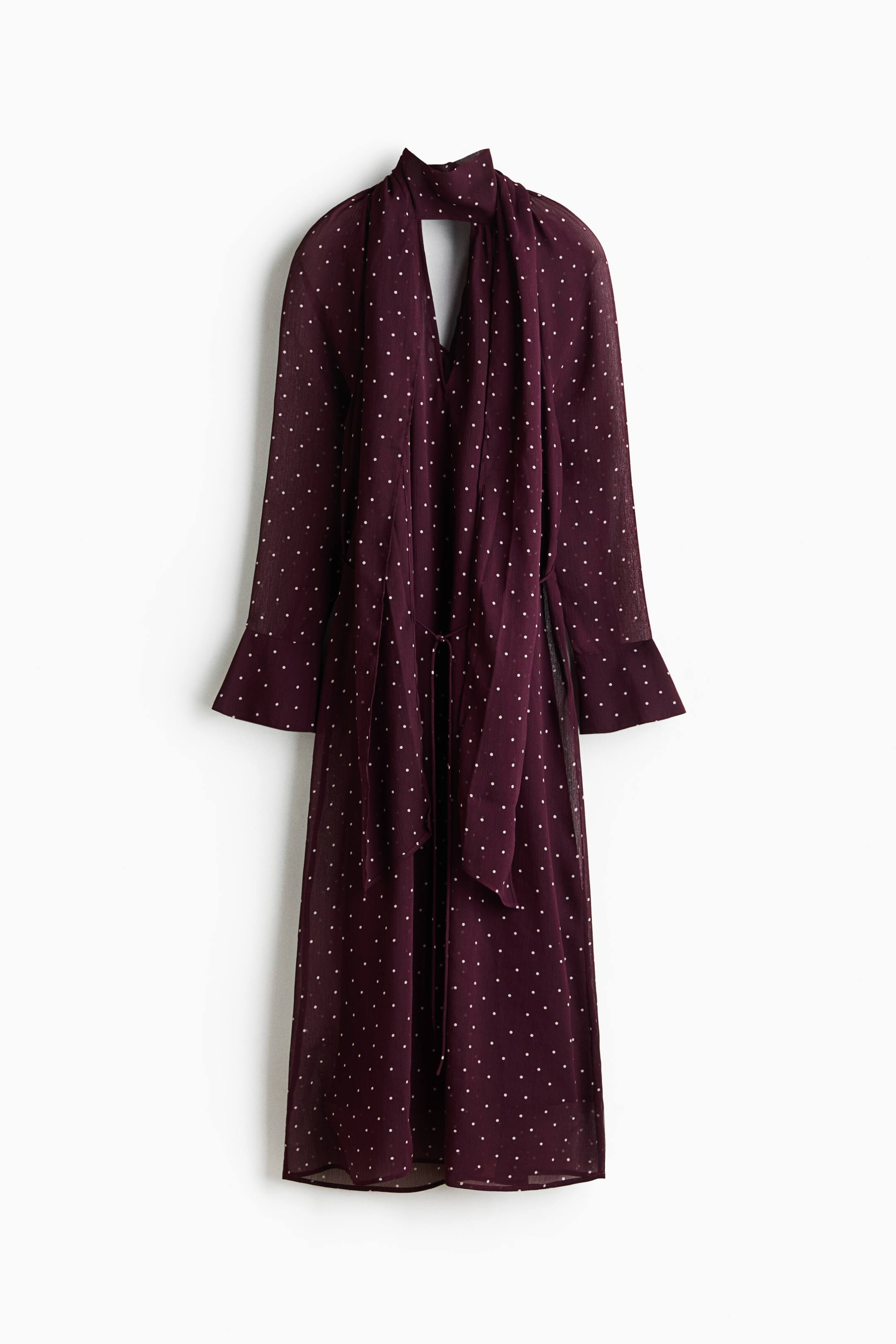 Ver imagen más grande: Un vestido midi burdeos, visto de frente, presenta un estampado de lunares blancos, cuello alto con largos lazos que crean una abertura en forma de V y una abertura tipo lágrima. También luce mangas largas con puños acampanados y un cinturón anudado del mismo tejido que ajusta la cintura.