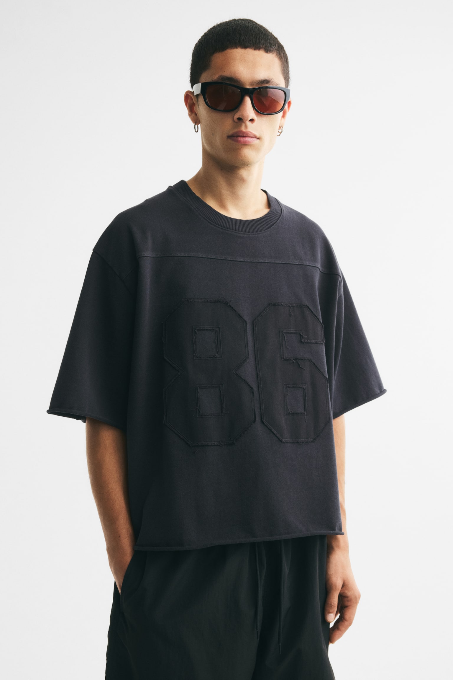 前面からディスプレイされたオーバーサイズの黒いコットンTシャツは、リブ付きのクルーネックと幅広のショートスリーブが特徴です。上部の胸部を横切る水平の縫い目があり、その下には大きな数字「86」がアップリケされており、微妙な質感のコントラストを演出しています。裾と袖の端は、未処理のエッジが見えています。 - 1
