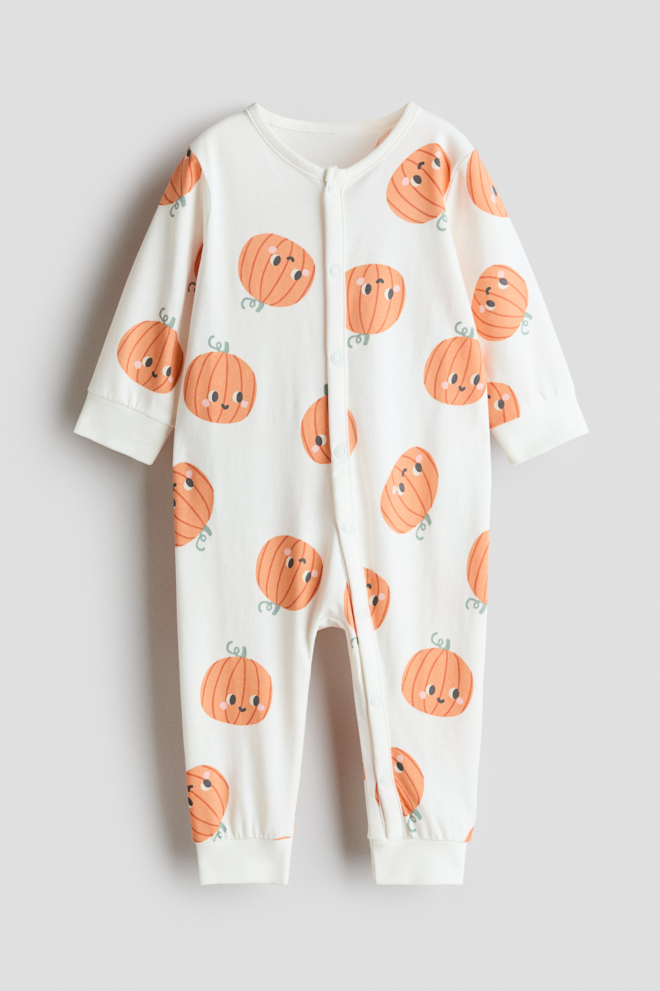 View larger image: An off-white cotton jersey sleepsuit is displaying an all-over print of smiling orange pumpkins, featuring long sleeves with plain cuffs and long legs with cuffed hems. Press-studs are running down the front and one leg.