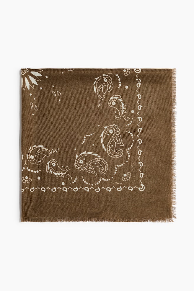 A large, square brown scarf is lying flat, facing front, showcasing a soft woven texture with a printed off-white paisley and floral design. The scarf has finely frayed edges along its borders. - 1