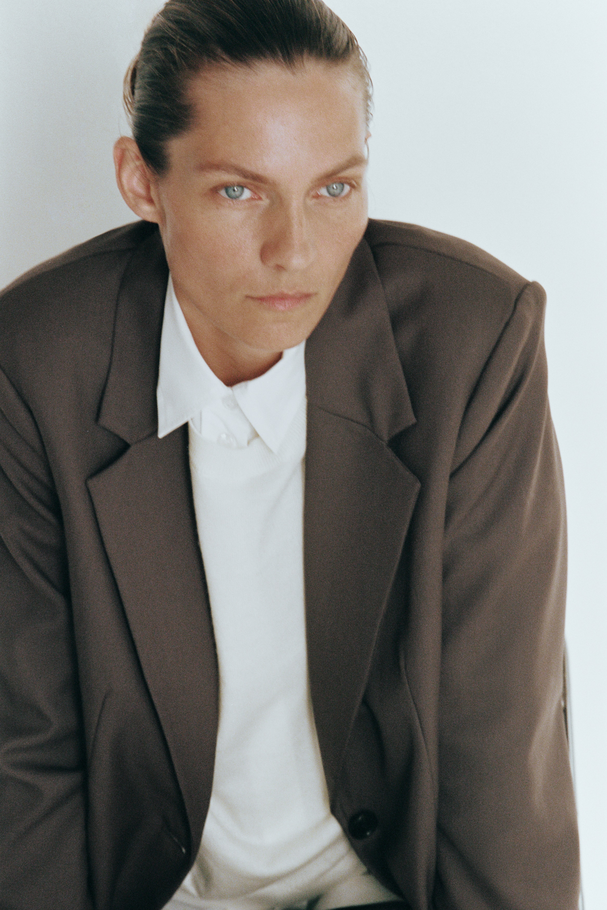 A single-breasted wool-blend blazer in taupe-brown is hanging front-facing, featuring wide notch lapels and a two-button closure. The blazer showcases structured shoulders, vertical waist darts, and two lower front pockets with flaps.