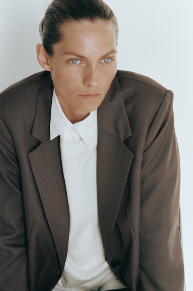 A dark brown, single-breasted blazer with notch lapels and a front button is being worn by the model. The blazer is layered over a white collared shirt and a cream-colored knit top. - 1