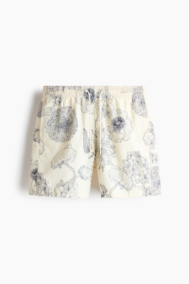 Swim shorts are presenting from the front, and are featuring a patterned weave in cream with a blue floral design. They are displaying a covered elasticated waist with a white drawstring, visible side pockets, and small slits at the side of the hems. - 1