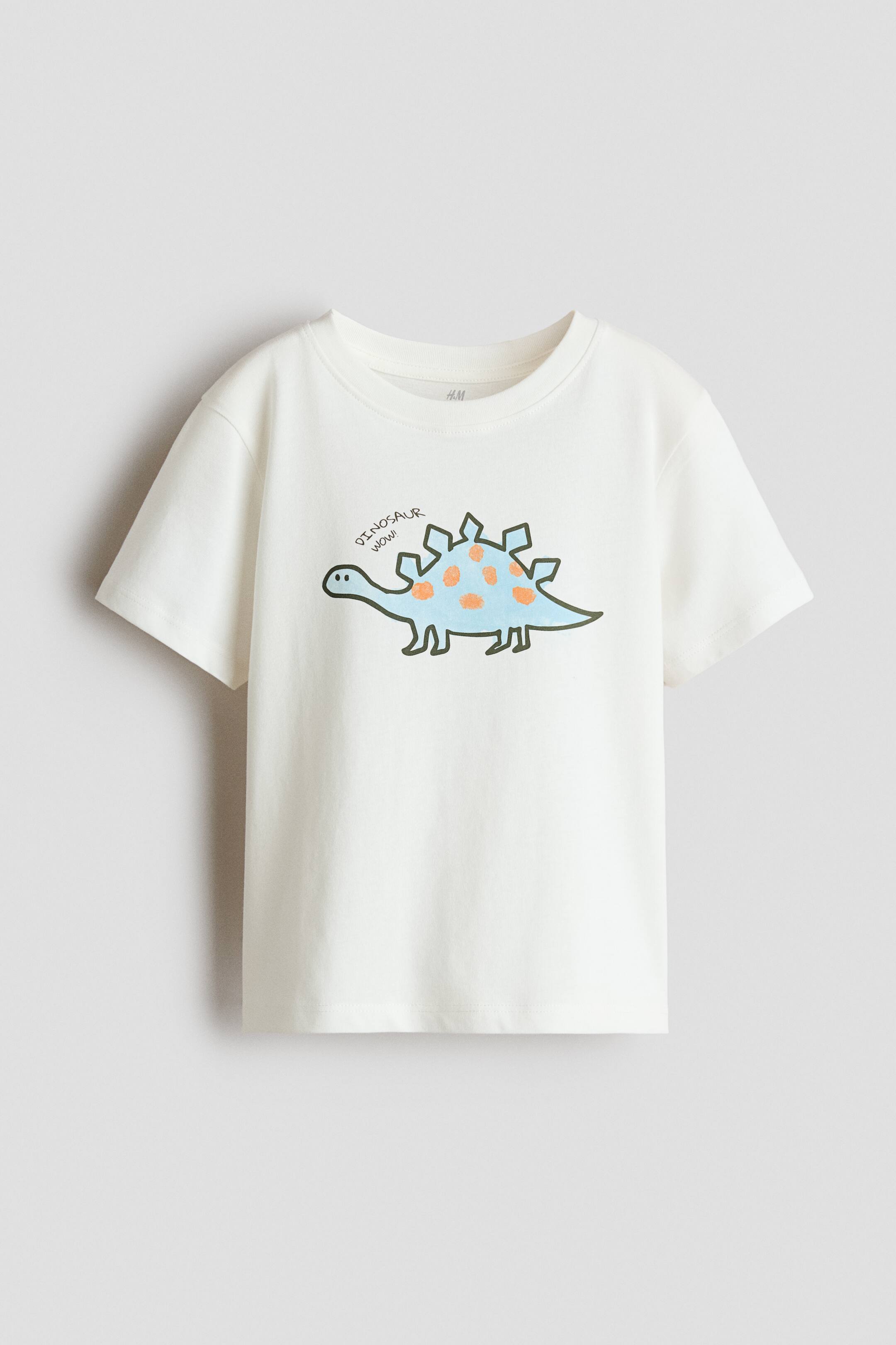 View larger image: A cream-coloured children's T-shirt is facing front, showcasing a classic crew neck and short sleeves. The centre features a cartoon graphic of a light blue stegosaurus with orange spots, with the text "DINOSAUR WOW!" printed in a playful font above it.