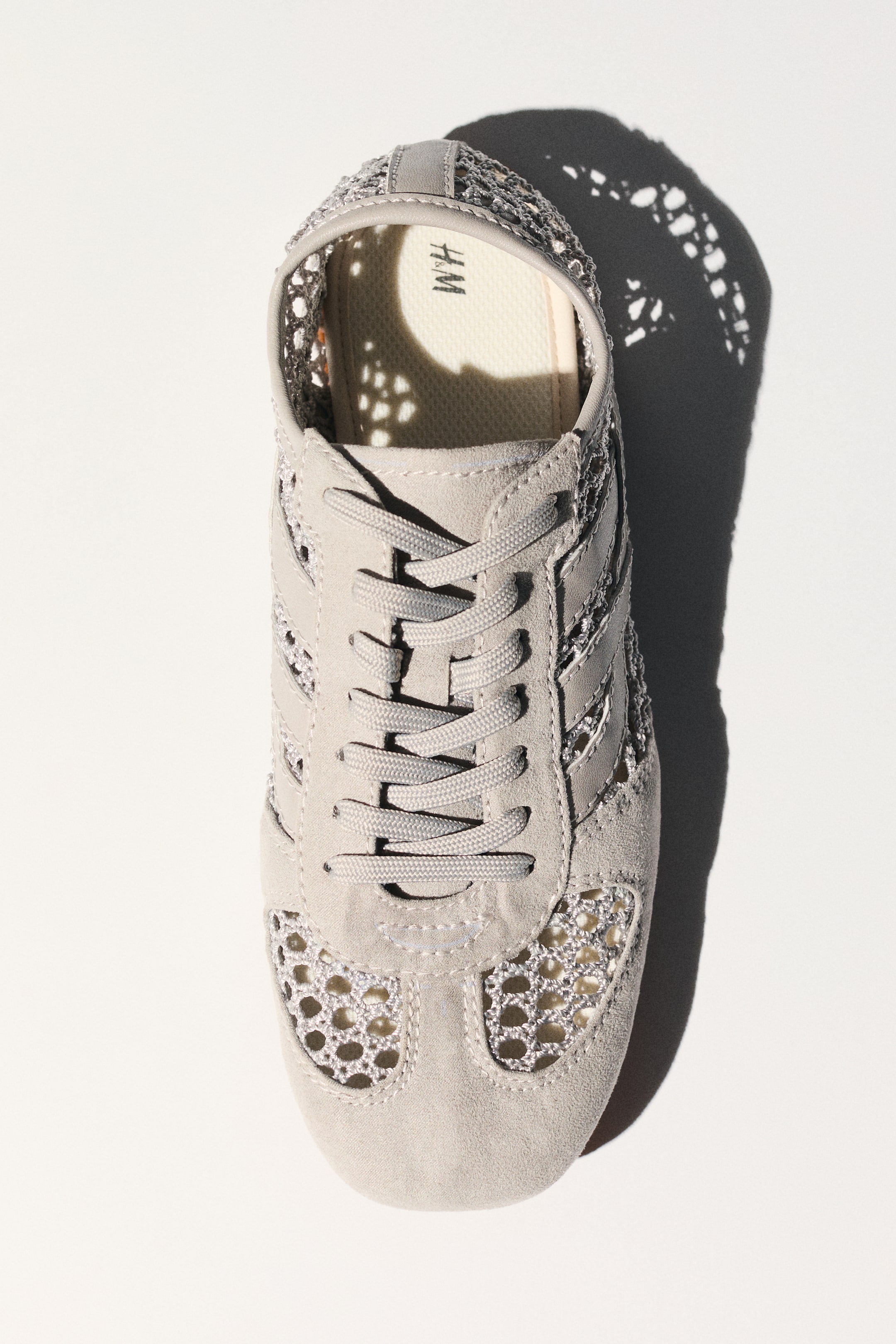 View larger image: A light gray sneaker is positioned facing forward, viewed from a top-down angle, highlighting a design with suede-like overlays and an open-knit mesh construction on the toe box and sides. It features a lace-up front with flat laces, and an 'H.M' logo is visible on the insole.