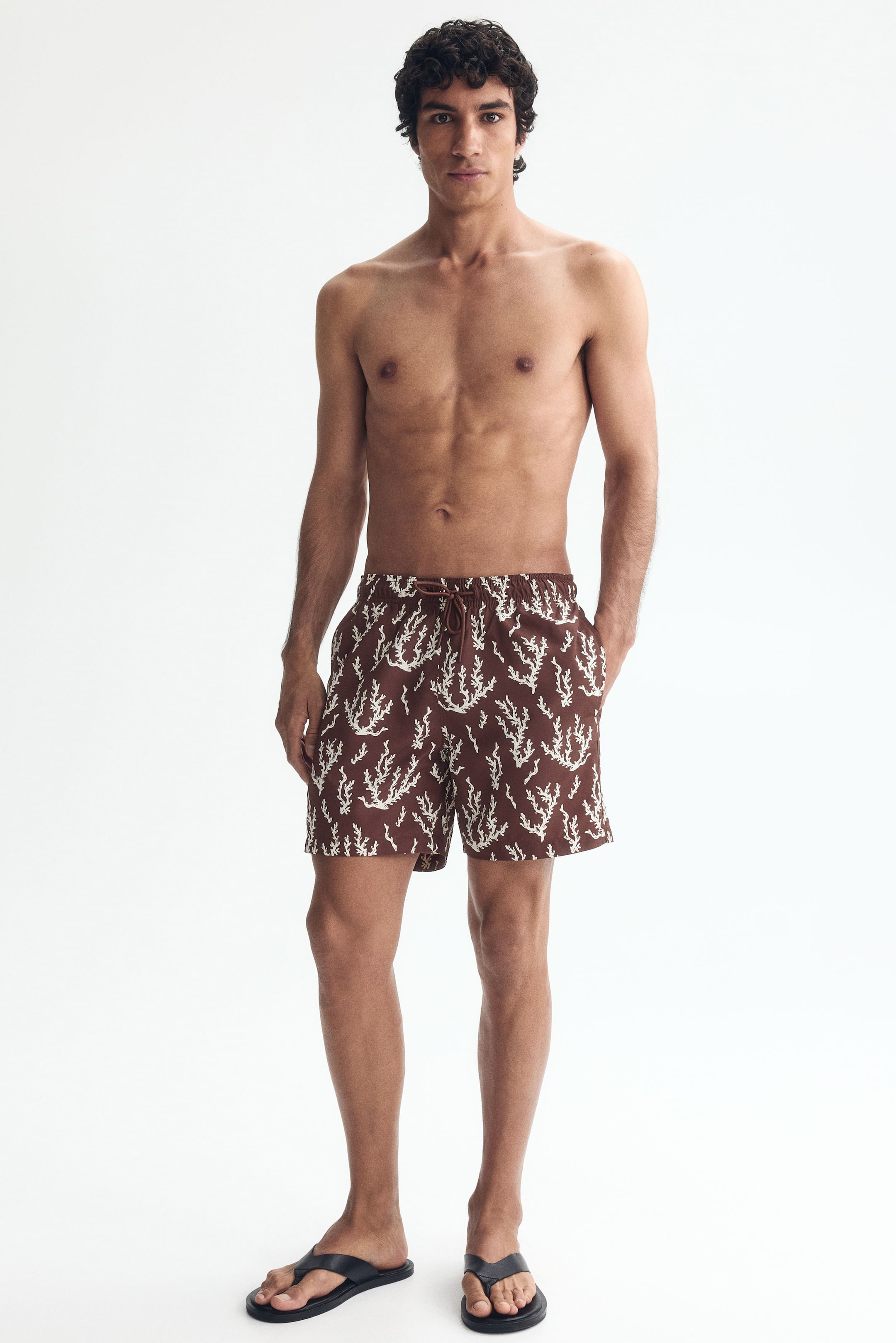 View larger image: The model is wearing swim shorts in a brown patterned weave, complete with an elasticated waist and drawstring. These shorts include side pockets and small slits at the side hems, with the model casually placing a hand in one pocket.