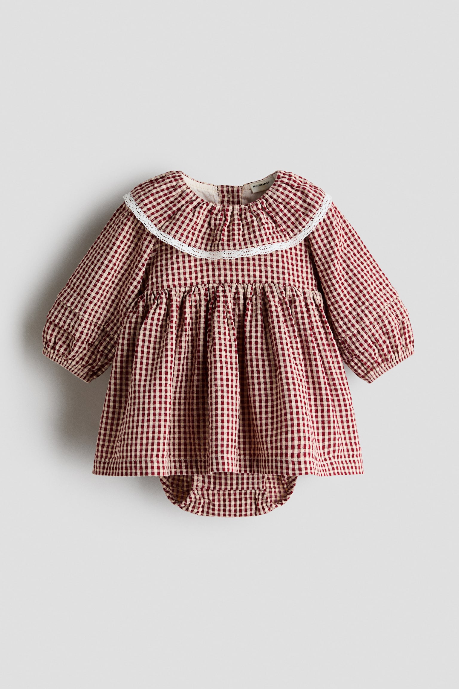 En babybodysuitklÃ¤nning i rÃ¶d- och vitrutigt gingham-mÃ¶nster ligger platt i framifrÃ¥nvy och har en bred volangkrage kantad med vit spets samt lÃ¥nga Ã¤rmar med resÃ¥r i Ã¤rmsluten. KlÃ¤nningen visar en rynkad hÃ¶g midja med utsvÃ¤ngd kjol och en matchande fastsydd bodysuit som skymtar under fÃ¥llen.