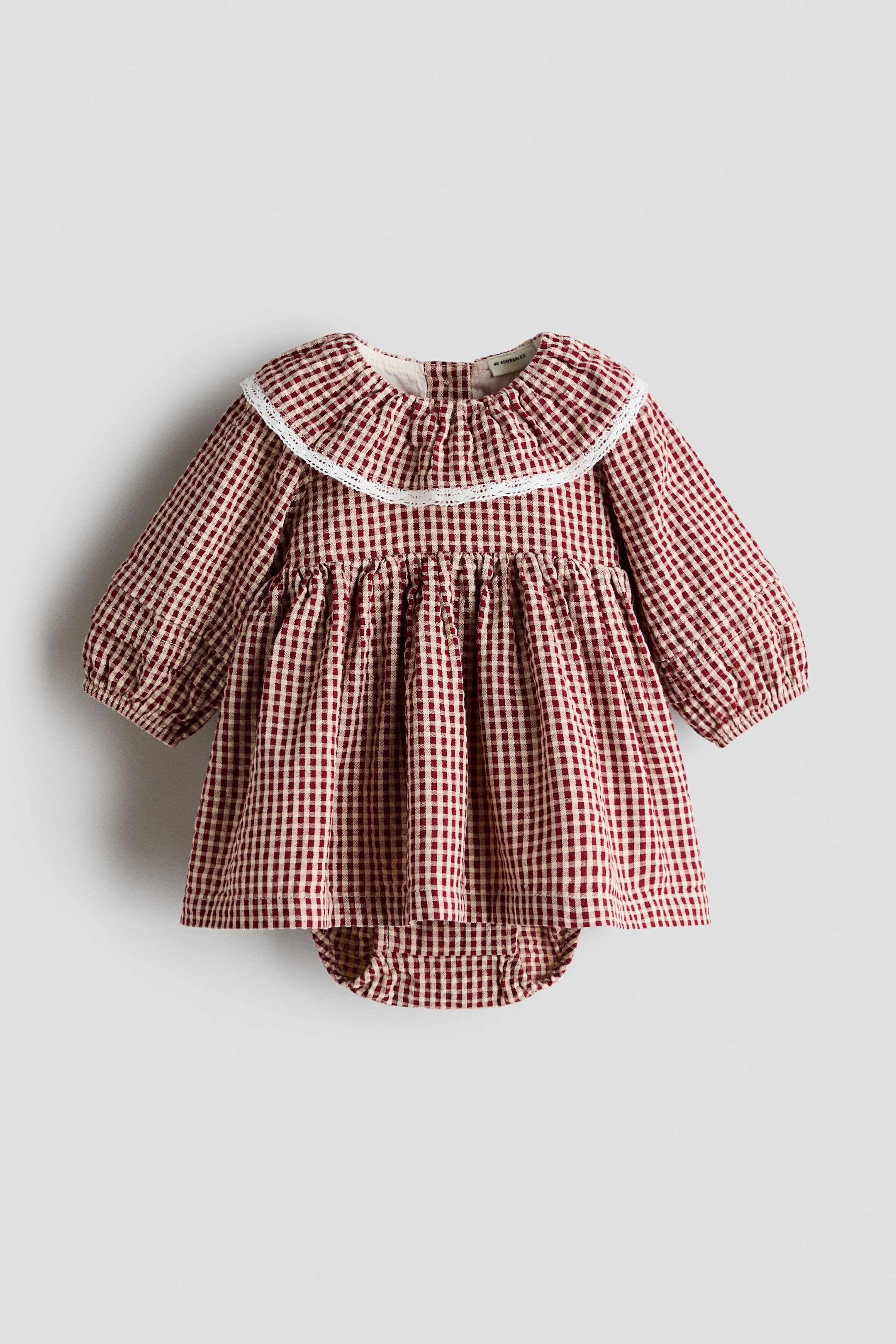 Agrandir l'image: Une robe-bodysuit pour bébé à motif vichy rouge et blanc est présentée à plat, vue de face, avec un large col volanté bordé de dentelle blanche et des manches longues à poignets élastiqués. La robe met en valeur une taille haute froncée avec une jupe évasée, ainsi qu’un bodysuit intégré assorti qui dépasse légèrement sous l’ourlet.