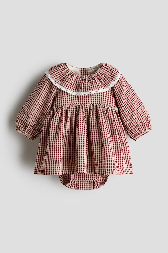 Punavalkoisella gingham-kuviolla oleva vauvan bodymekko on aseteltu tasaisesti eteenpÃ¤in suunnattuna kuvana. SiinÃ¤ on leveÃ¤, valkoisella pitsillÃ¤ viimeistelty rÃ¶yhelÃ¶kaulus sekÃ¤ pitkÃ¤t hihat resoreilla varustetuilla manseteilla. Mekossa on poimutettu korkea vyÃ¶tÃ¤rÃ¶, levenevÃ¤ hameosa ja helman alta pilkistÃ¤vÃ¤ yhteensopiva kiinniommeltu bodyosa. - 1