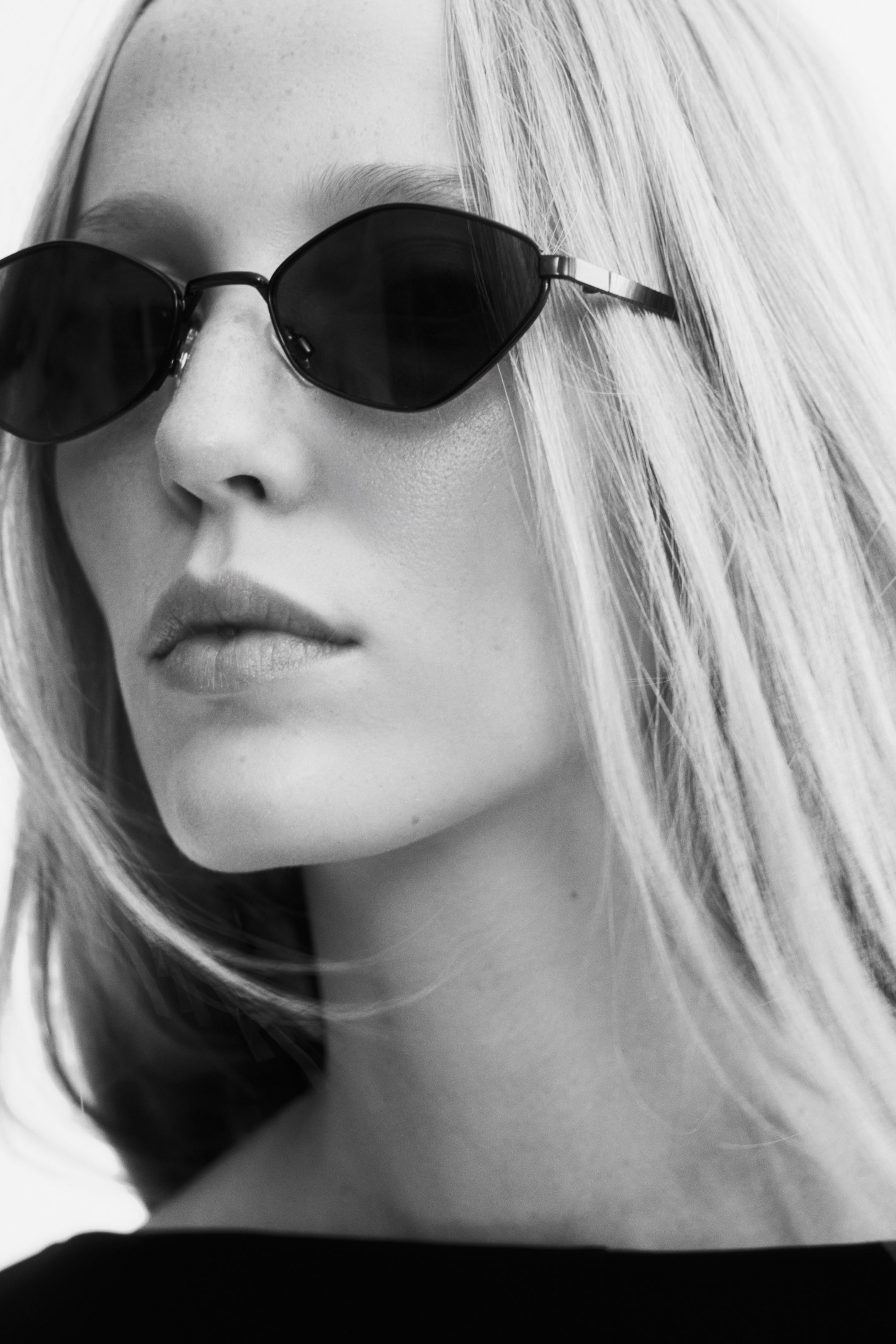 View larger image: Geometric oval sunglasses with thin metal frames and dark tinted lenses are worn by the model. The close-up view highlights the angular design of the frames and the slender metal temples.