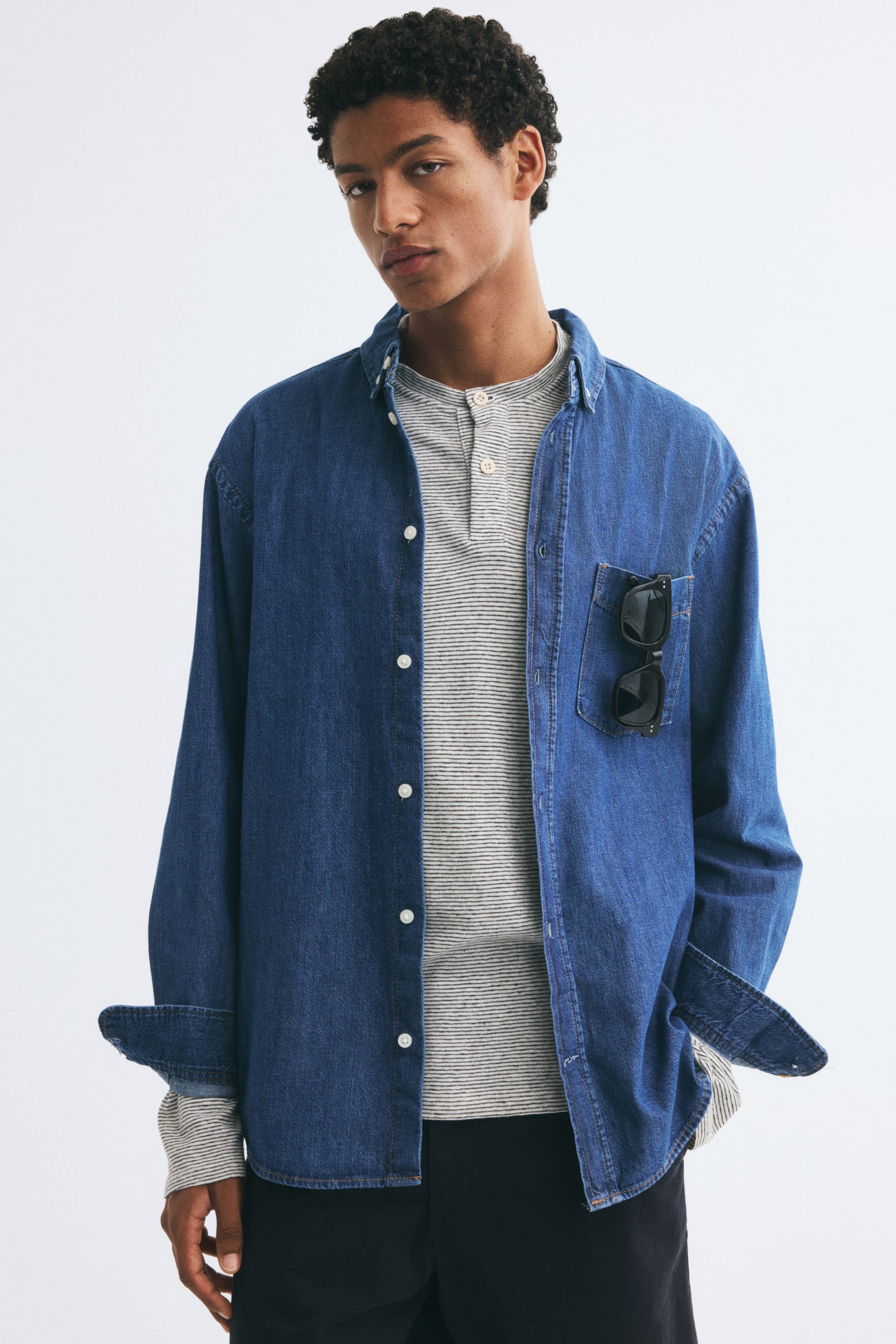 Se større billede: En blå denimskjorte bæres åben af modellen, lagt over en stribet henley-trøje og sorte bukser. Skjorten har klassisk krave, hvide knapper og en påsyet brystlomme, der rummer et par sorte solbriller.