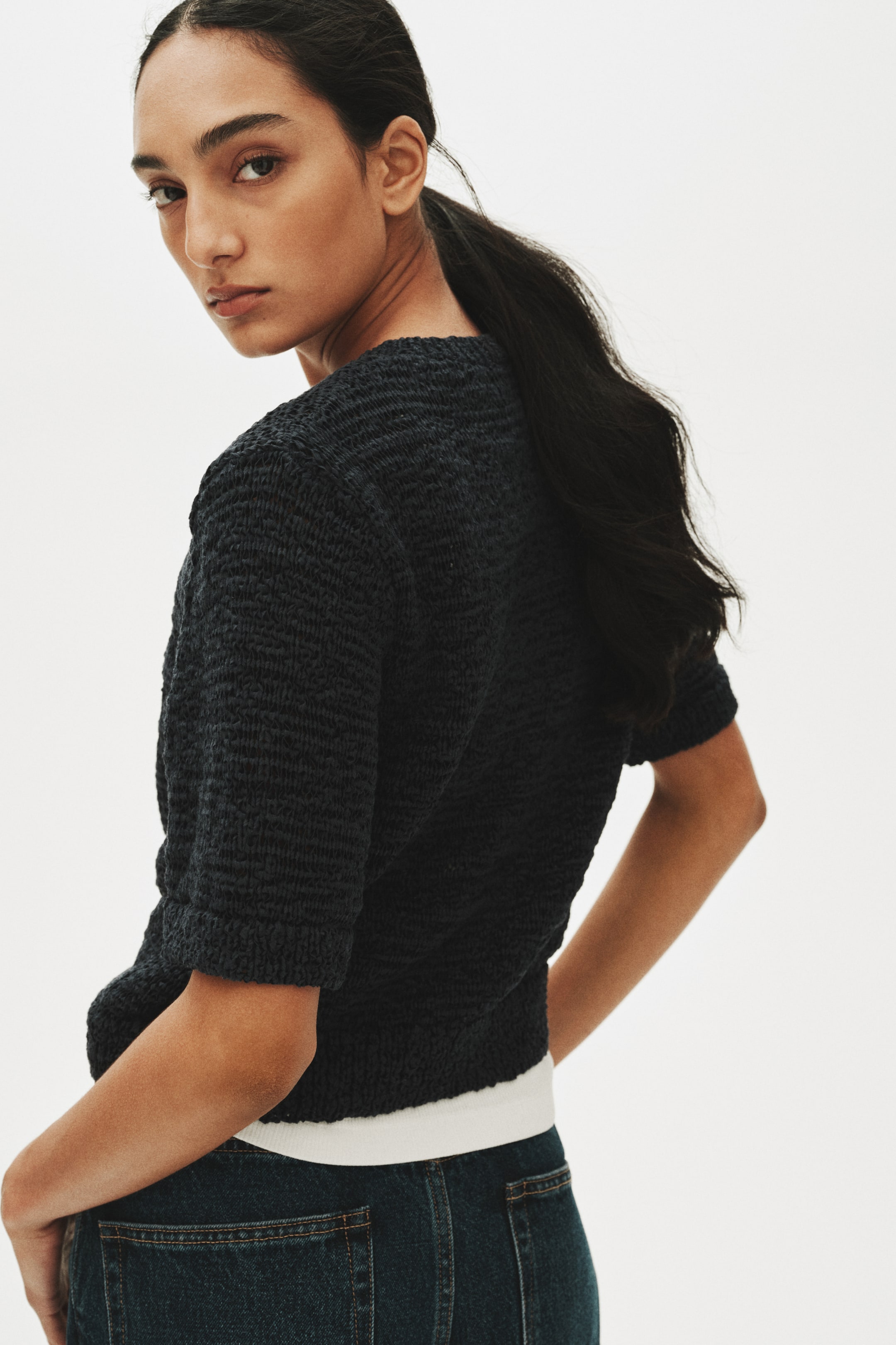 View larger image: A dark charcoal textured knit top is shown from the back and side, featuring short sleeves with cuffed hems. The top displays a distinct, raised knit pattern across the fabric and finishes at the waist, revealing a white layer underneath.