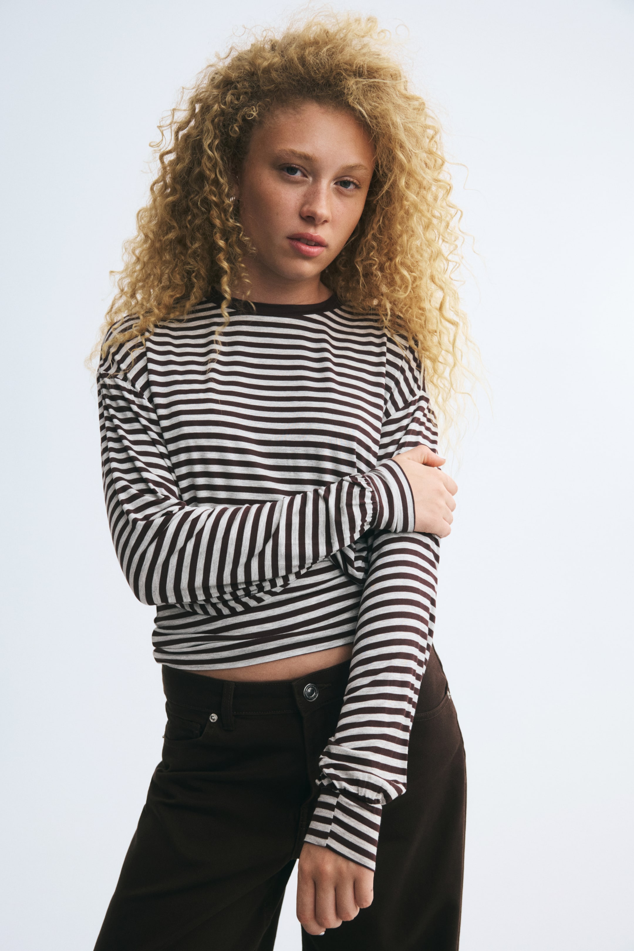 View larger image: The model is wearing a long-sleeved crewneck T-shirt with horizontal brown and off-white stripes. They are posing with one arm folded across the body, holding the upper arm, and styling the top with dark brown pants.