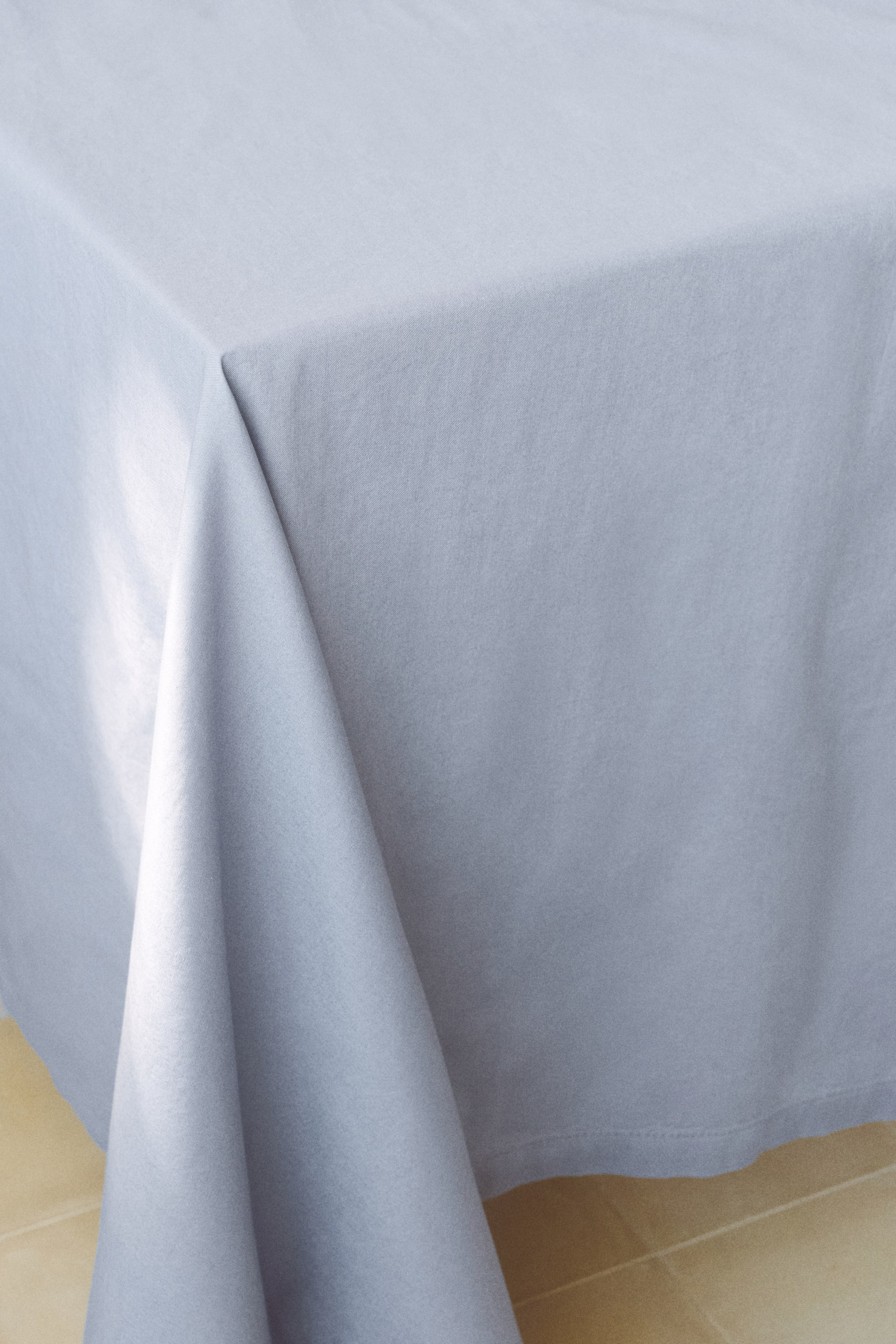 View larger image: A light gray tablecloth in cotton canvas is draped over a table, showing its front corner. It features hemstitch detailing and a 1-inch hem along its edges.