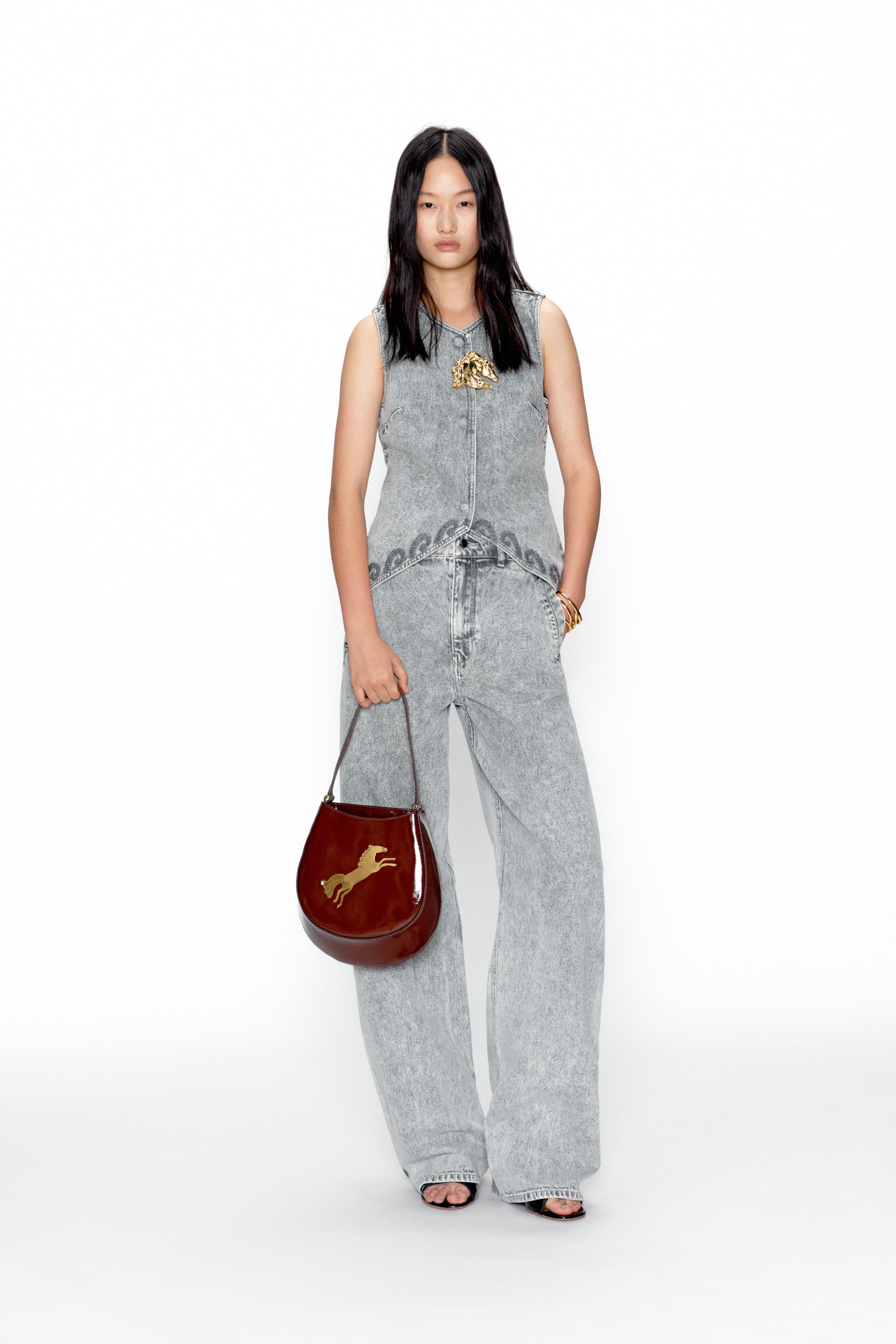 View larger image: A coordinated grey acid-wash denim vest and wide-leg jeans set is shown from the front. The vest has a V-neckline, front buttons, and a gold-tone ornate brooch, while both pieces feature a subtle swirling pattern around the waist. A glossy reddish-brown crescent-shaped bag with a golden rearing horse emblem is carried to complement the outfit.