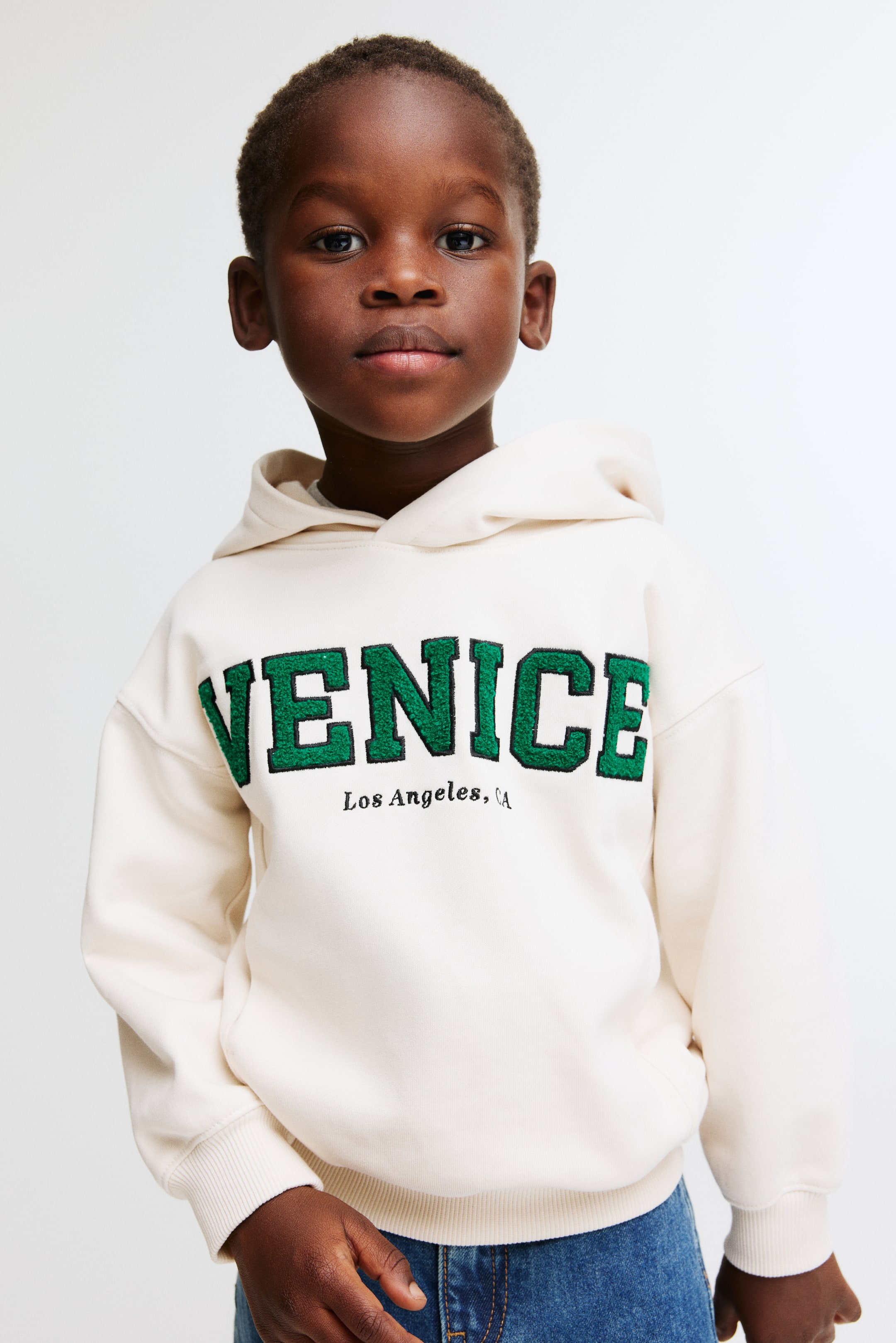 View larger image: The cream-colored hoodie is facing forward, displaying a large green embroidered "VENICE" text with a black outline on its front, along with "Los Angeles, CA" embroidered beneath it. It is also featuring a hood, ribbed cuffs, a ribbed hem, and a front kangaroo pocket.