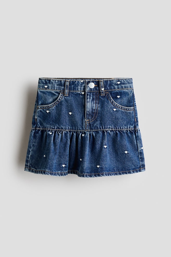 Ein blauer Jeansrock wird von vorne präsentiert, mit einem Bund mit Gürtelschlaufen, einem silberfarbenen Knopf, einem Reißverschluss und vorderen Schaufeltaschen. Kleine weiße Herzmuster sind über den Jeansstoff verstreut, der auch eine geraffte Rüschenschicht am unteren Saum enthält. - 1