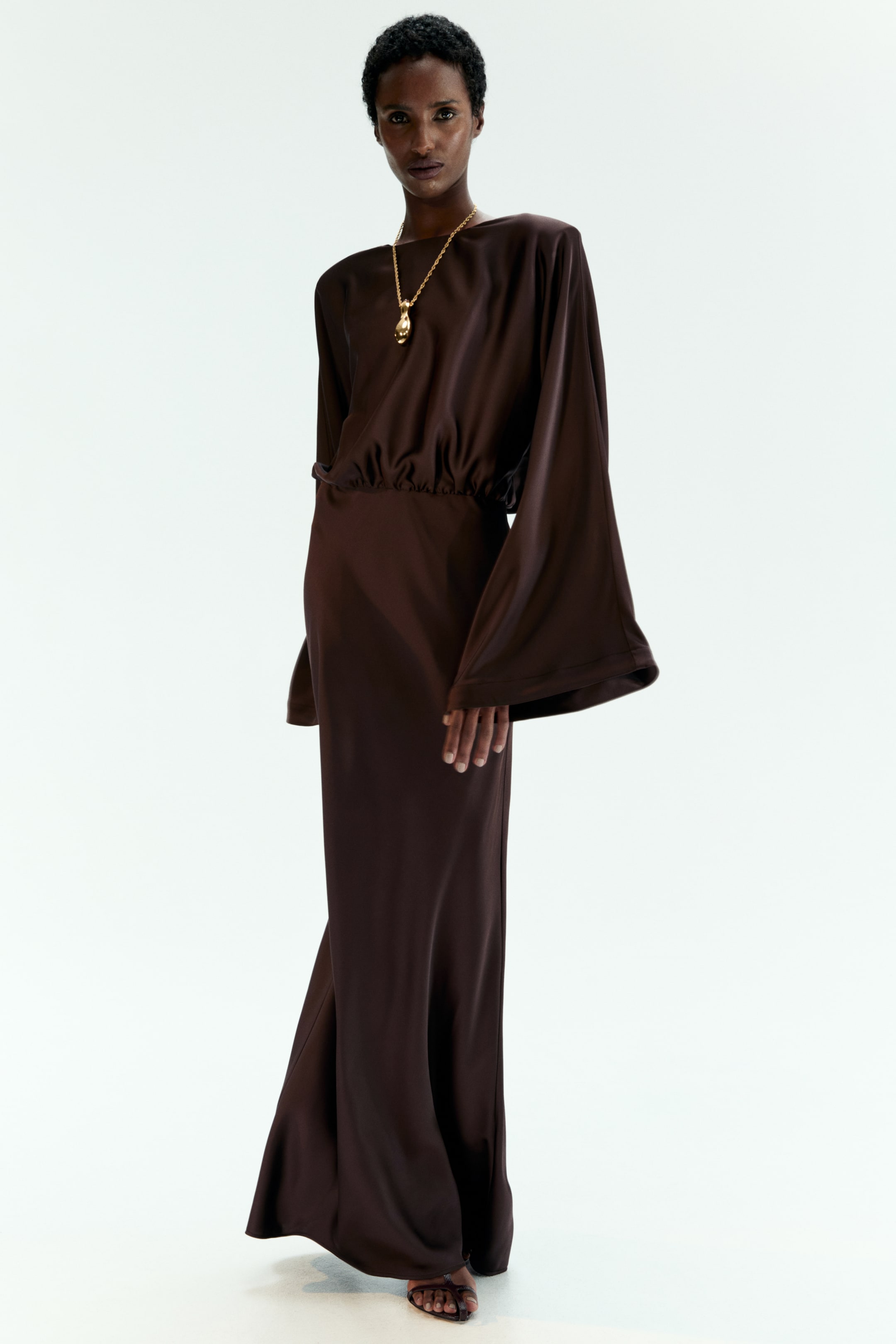 Visa större bild: En brun maxiklänning med båthalsringning, strukturerade axlar, vida ärmar och en rynkad midja bärs av modellen. Den flödande, golvlånga klänningen är stylad med ett guldhänge och mörka sandaler.
