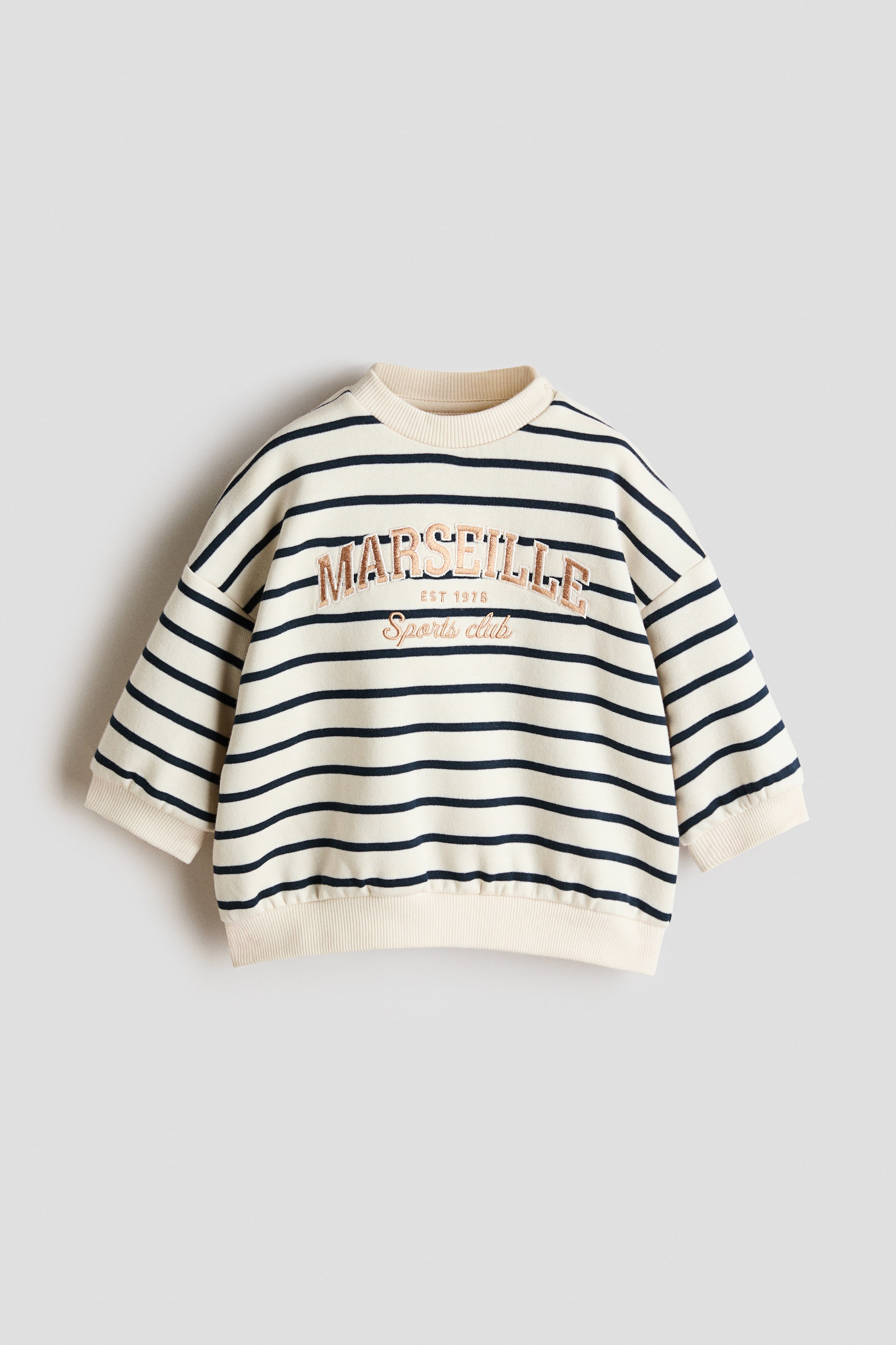 View larger image: A front-facing cream sweatshirt with navy horizontal stripes features "MARSEILLE EST 1978 Sports club" embroidered in bronze-toned thread on the chest. It's designed with a ribbed crewneck, ribbed cuffs, and a ribbed hem.