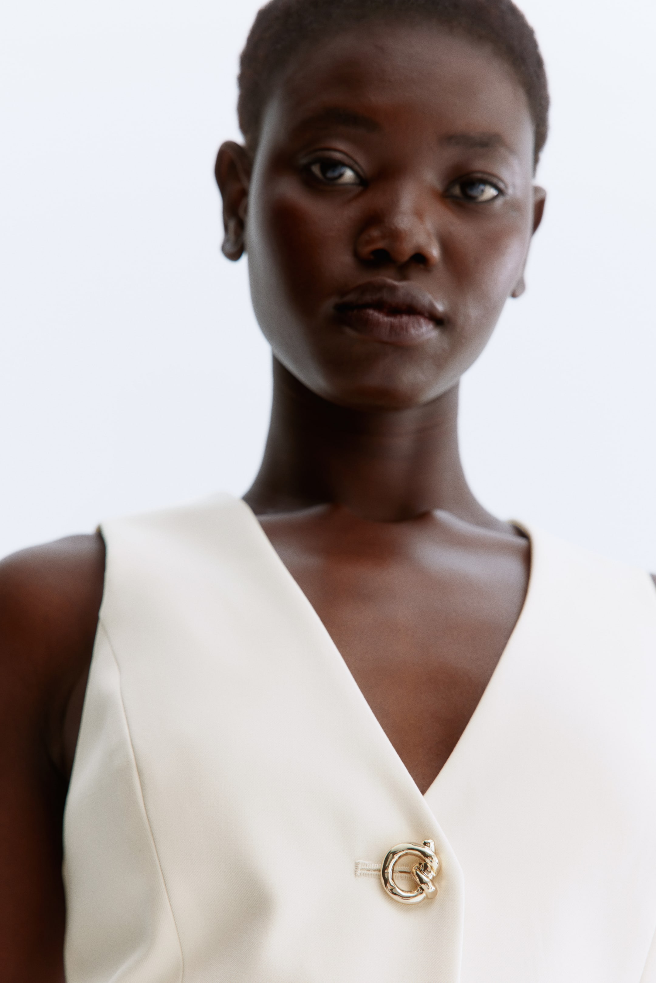 Ampliar la imagen: Un chaleco sin mangas de color crema presenta un escote en V y un destacado broche circular en tono dorado que cierra el frente.