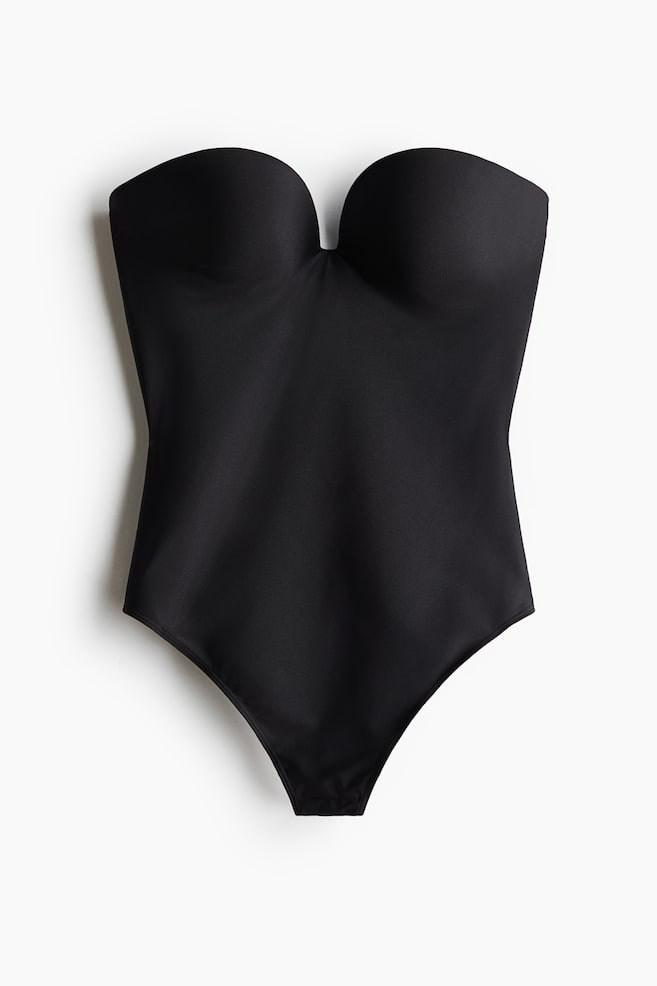 A black shaping bodysuit is laid flat in a front view, showcasing its seamless microfibre construction. It features structured, moulded cups with a sweetheart neckline and high-cut leg openings. - 1