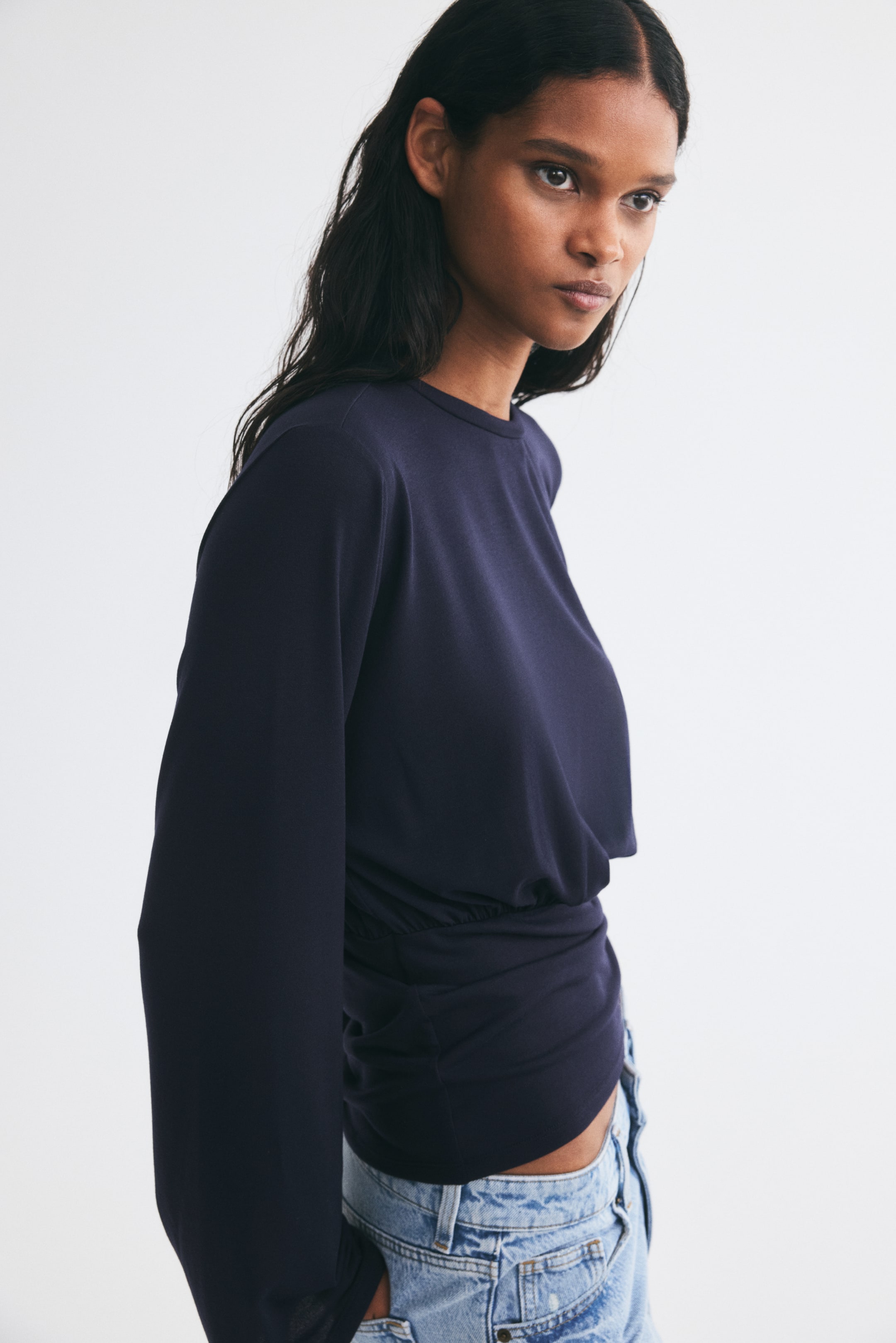 View larger image: The model is wearing a dark blue long-sleeved top. The top features a crew neck and a gathered waist, styled with light wash denim jeans.