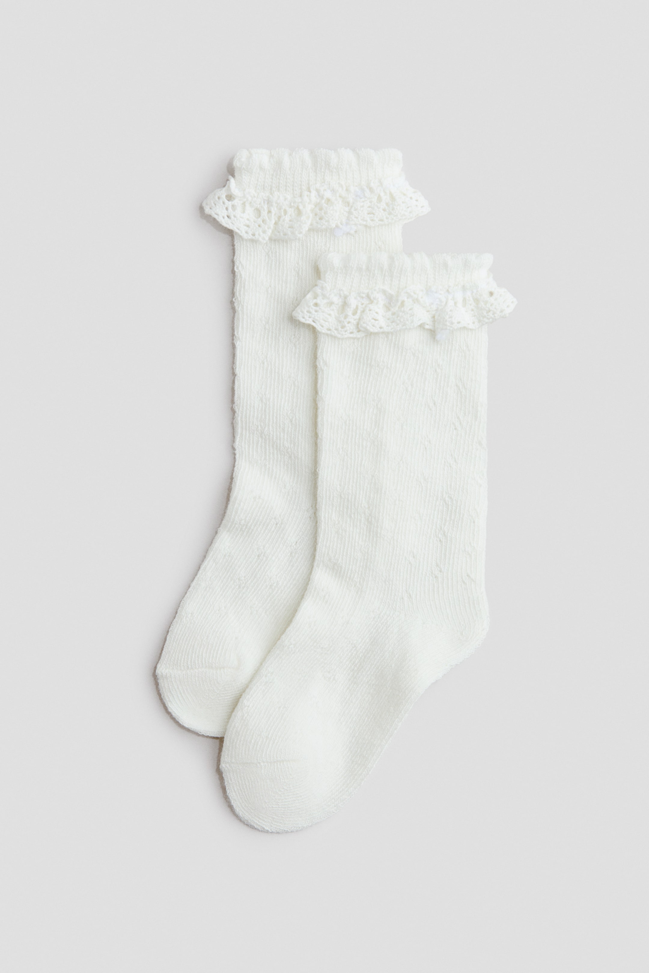Prikaži veću sliku: Predstavljamo par belih dokolenica, izrađenih od mekog, finog pletiva sa suptilnim teksturiranim uzorkom. Svaka čarapa ima elastičnu manžetnu, ukrašenu naboranom čipkom i malom mašnicom.