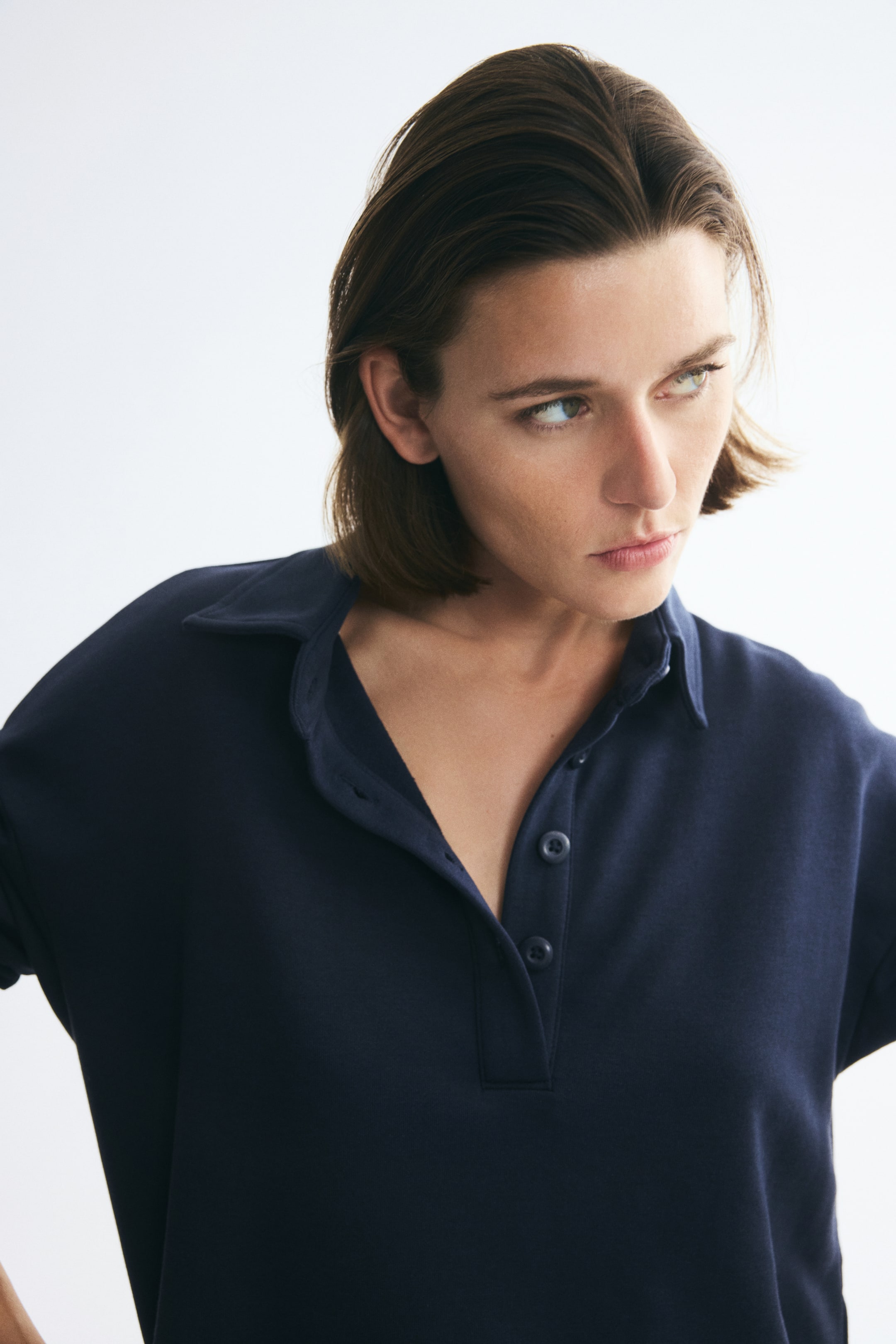 Agrandir l'image: Un pull bleu foncé en tissu sweat léger est porté par le mannequin, avec un col plat et une patte de boutonnage partiellement ouverte. La vue en gros plan met en valeur la coupe décontractée et le design à épaules tombantes du vêtement.