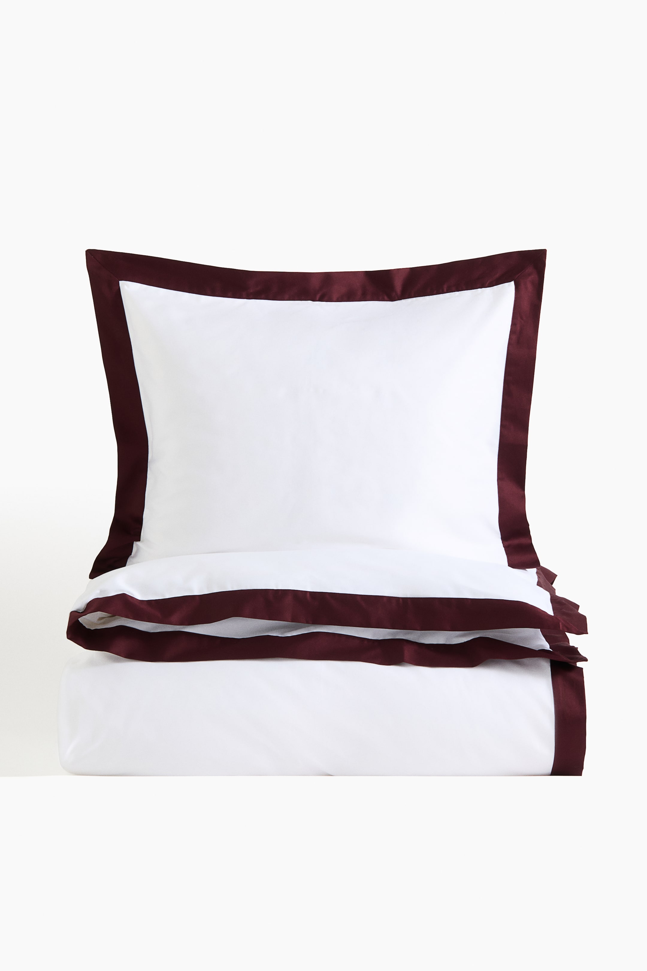 Agrandir l'image: Un ensemble de linge de lit blanc est présenté, comprenant une taie d’oreiller et une housse de duvet pliée. Les deux pièces présentent un large passepoil satiné bordeaux bordant leurs contours. La taie d’oreiller est dressée verticalement, légèrement inclinée vers la droite, au-dessus de la housse de duvet pliée à l’horizontale.