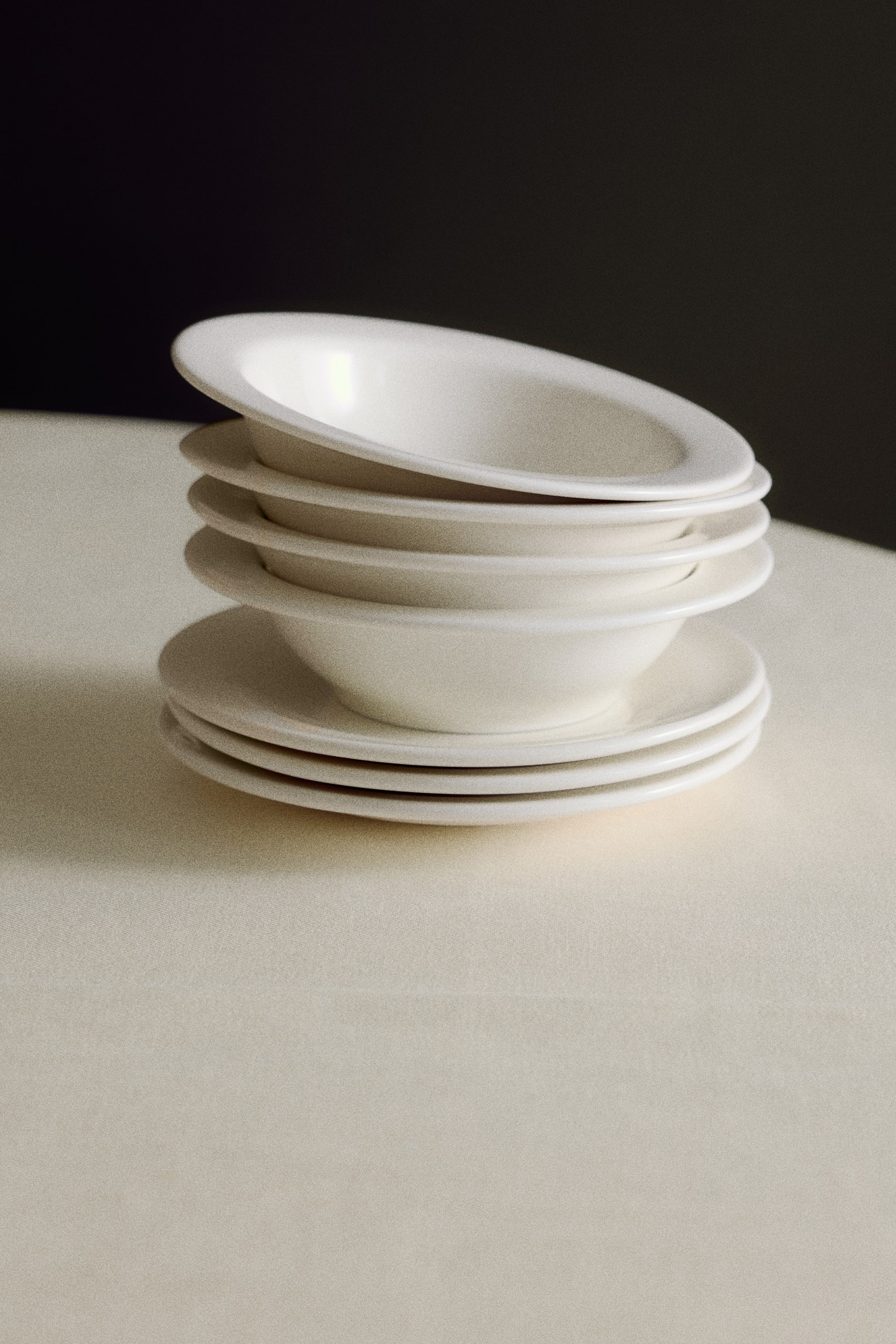 View larger image: Four small porcelain bowls with a minimalist design and a classic wide rim are stacked on top of three matching plates, with the top bowl tilted slightly to show its shallow, rounded basin.