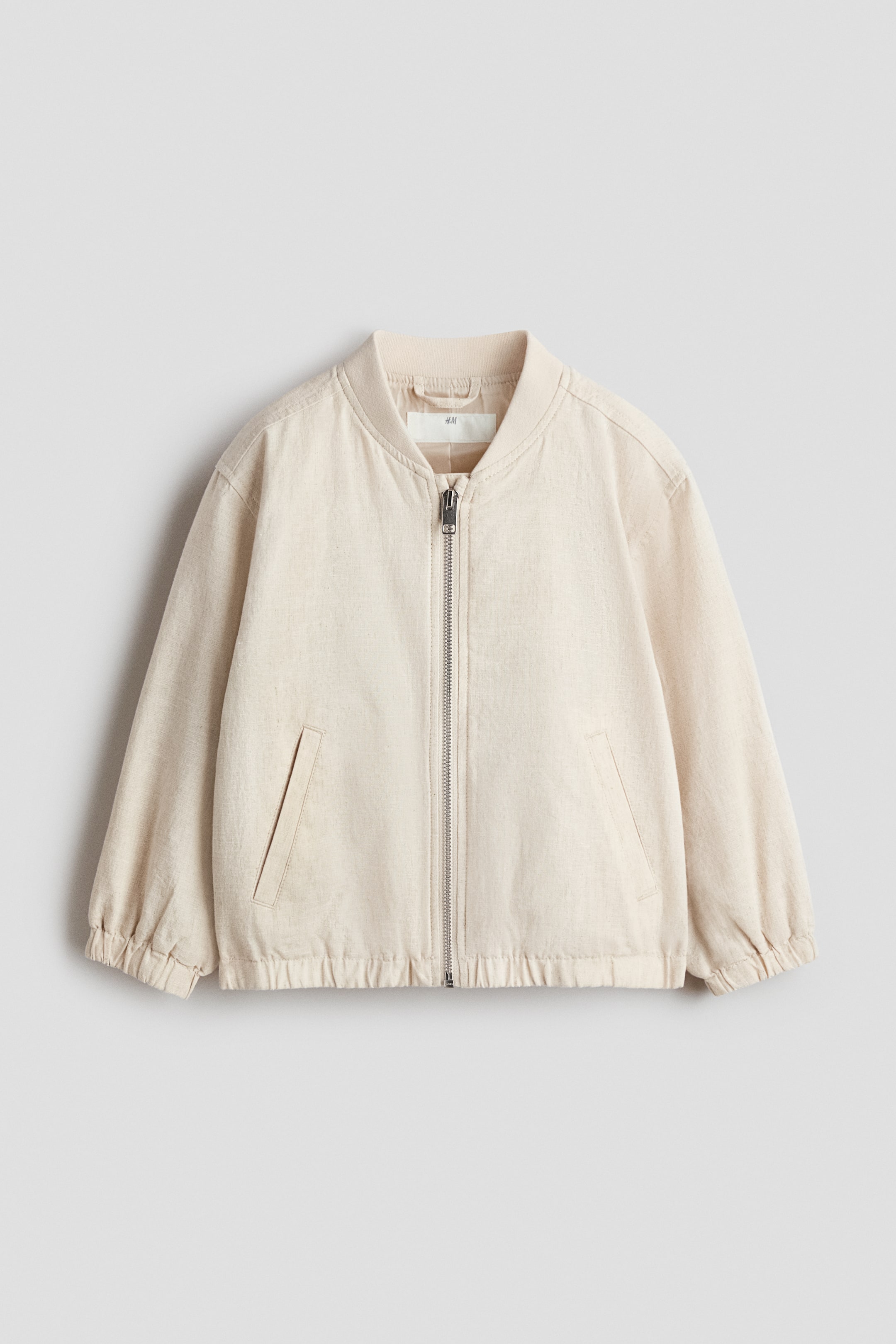 Ver imagen más grande: Una chaqueta bomber de color beige claro presenta su parte frontal, con un cuello alto acanalado, una cremallera frontal en tono plateado y dos bolsillos inclinados de ribete. También muestra puños elásticos y un dobladillo elástico.