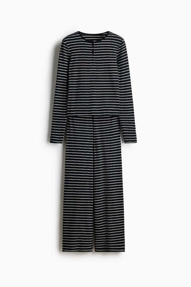 Una vista frontal muestra un conjunto de pijama de punto pointelle texturizado con rayas horizontales en blanco y negro. El conjunto incluye una parte superior de manga larga con cuello redondo y tapeta de botones, combinada con un pantalón recto a juego. - 1