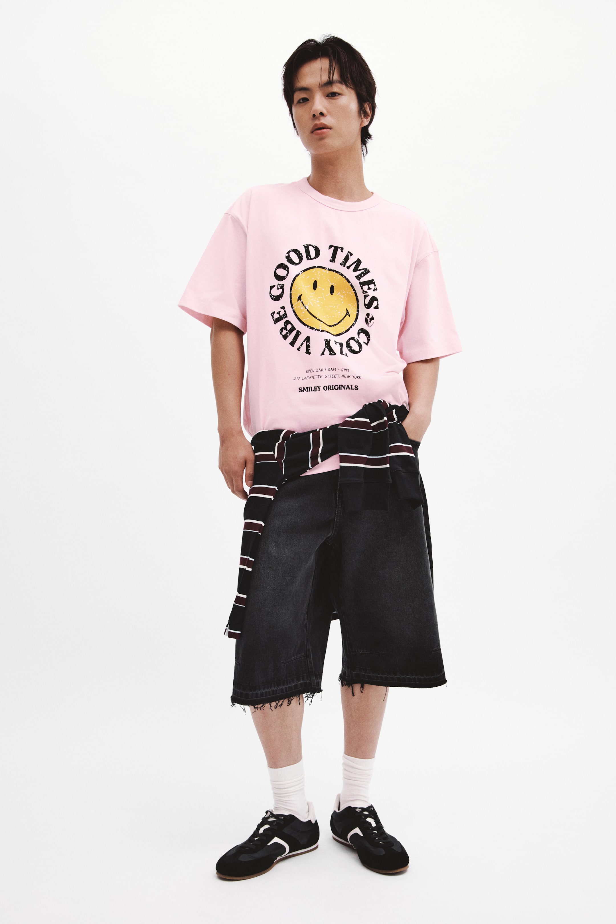 Xem ảnh lớn hơn: Một chiếc áo phông màu hồng bằng vải jersey cotton dày in hình đang được người mẫu mặc, nổi bật với đồ họa mặt cười màu vàng lớn ở phía trước. Áo có cổ tròn viền gân, vai rơi và dáng rộng, được sơ vin một phần vào quần short.