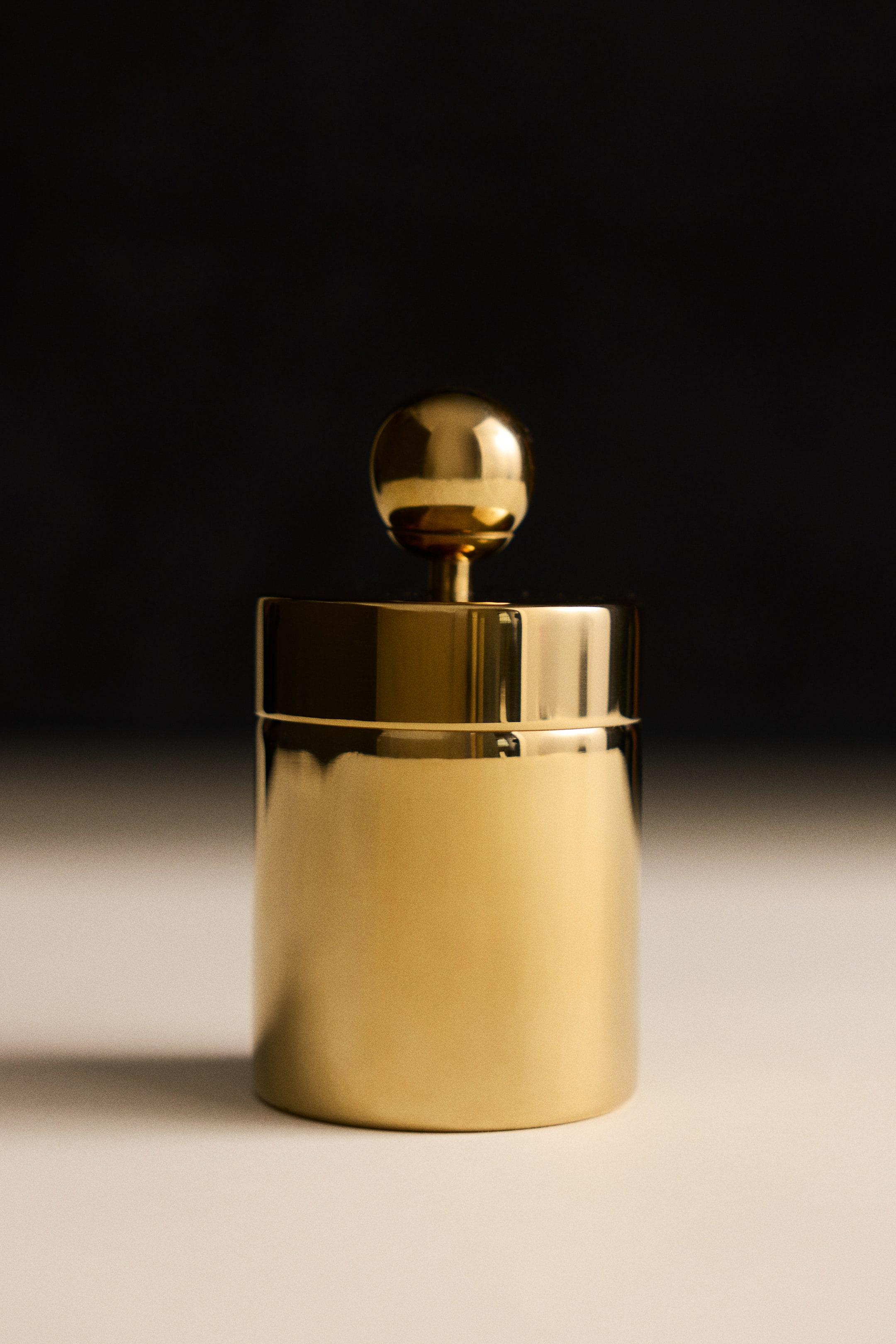 View larger image: A tall, round metal canister with a polished gold finish is standing upright, featuring a cylindrical body and a matching lid. The lid has a prominent spherical knob on a short, central stem.
